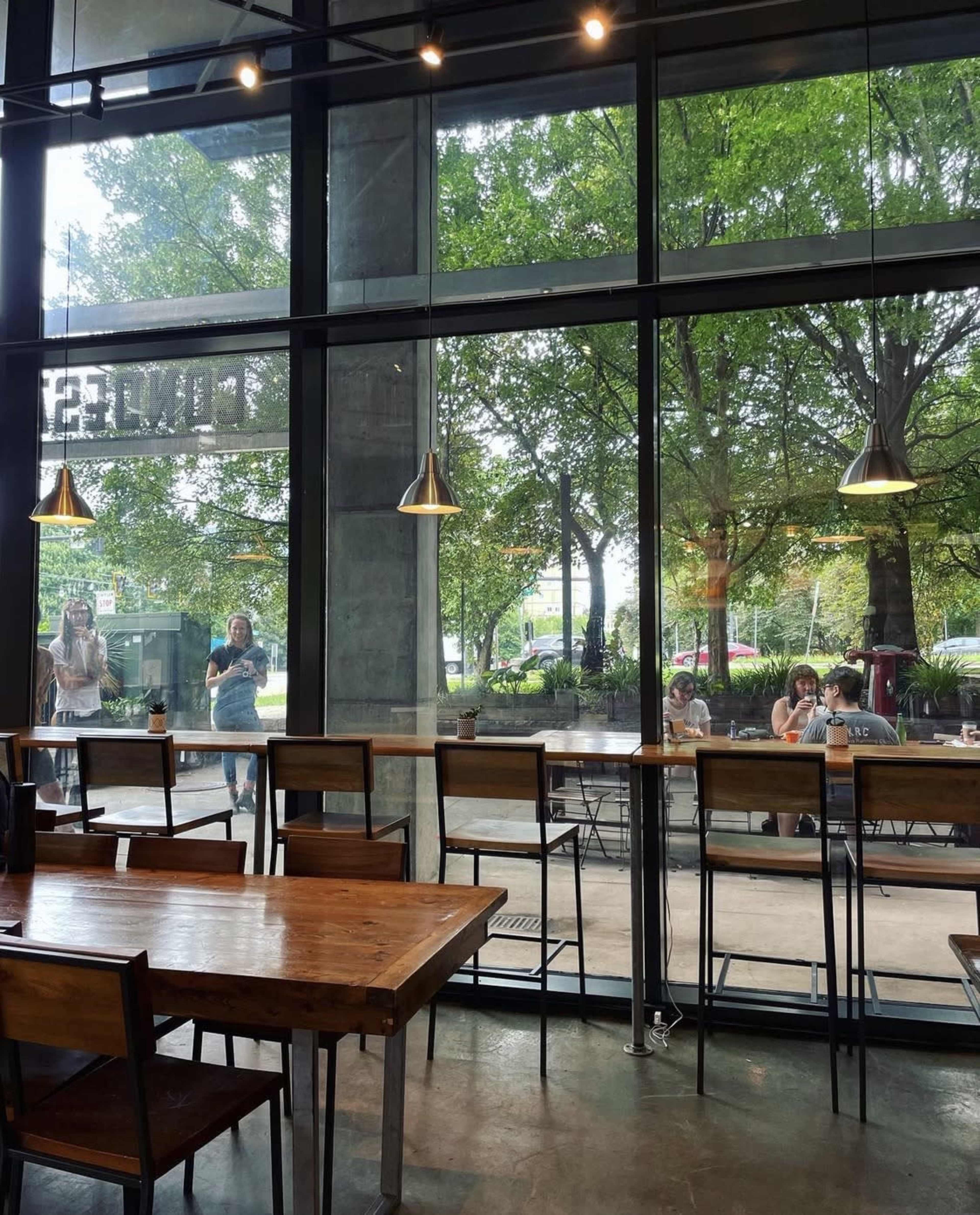 The image shows a modern café interior with wooden tables and chairs, large windows providing a view of people outside, and greenery surrounding the area.