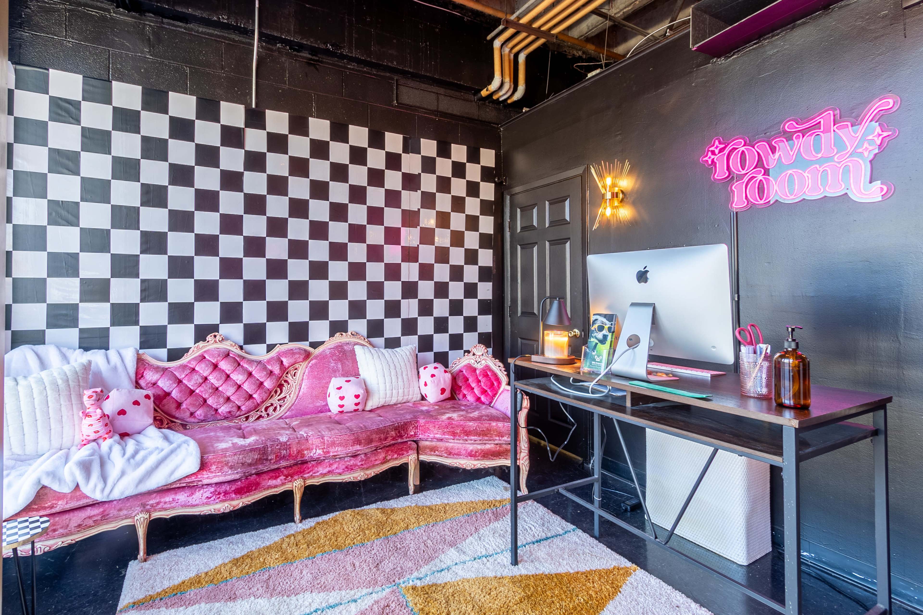 A small room featuring a pink, tufted couch, a checkered wall, and a desk with a computer.