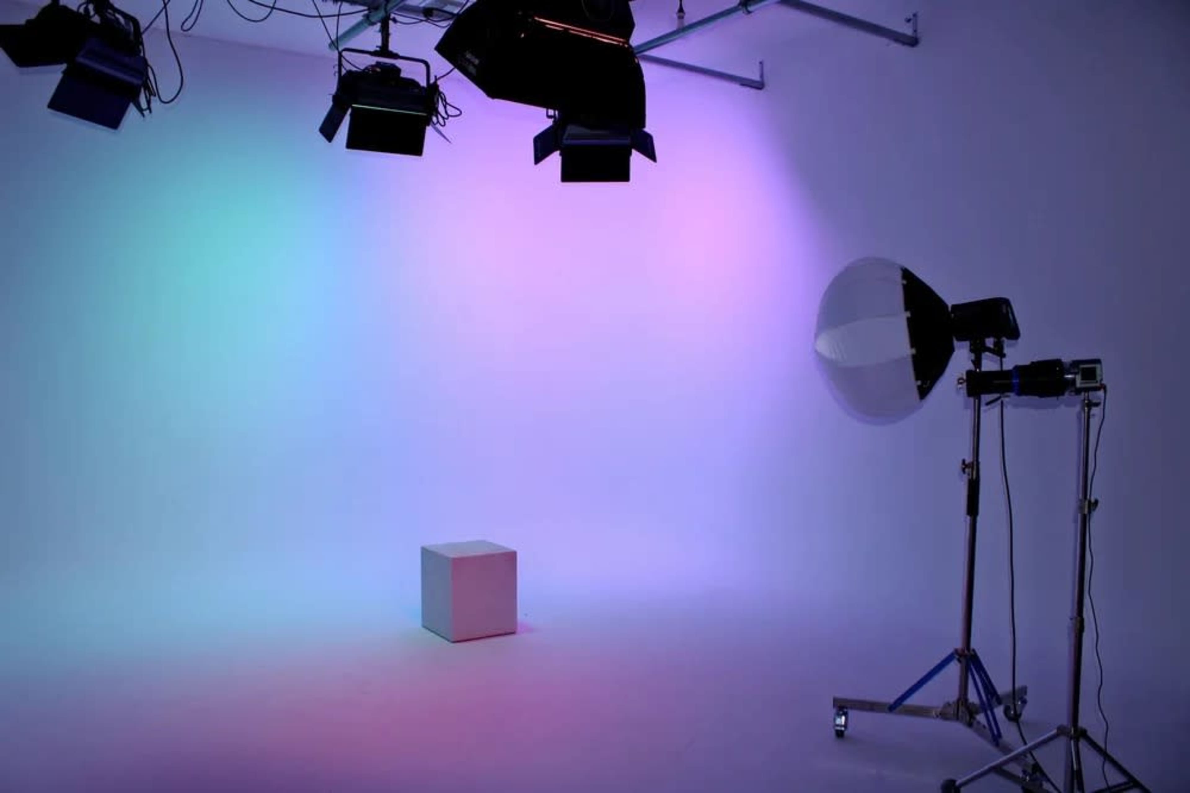 The image shows a studio setting with colorful lights illuminating a white cube on the floor and a photography setup including a large reflector on a stand.