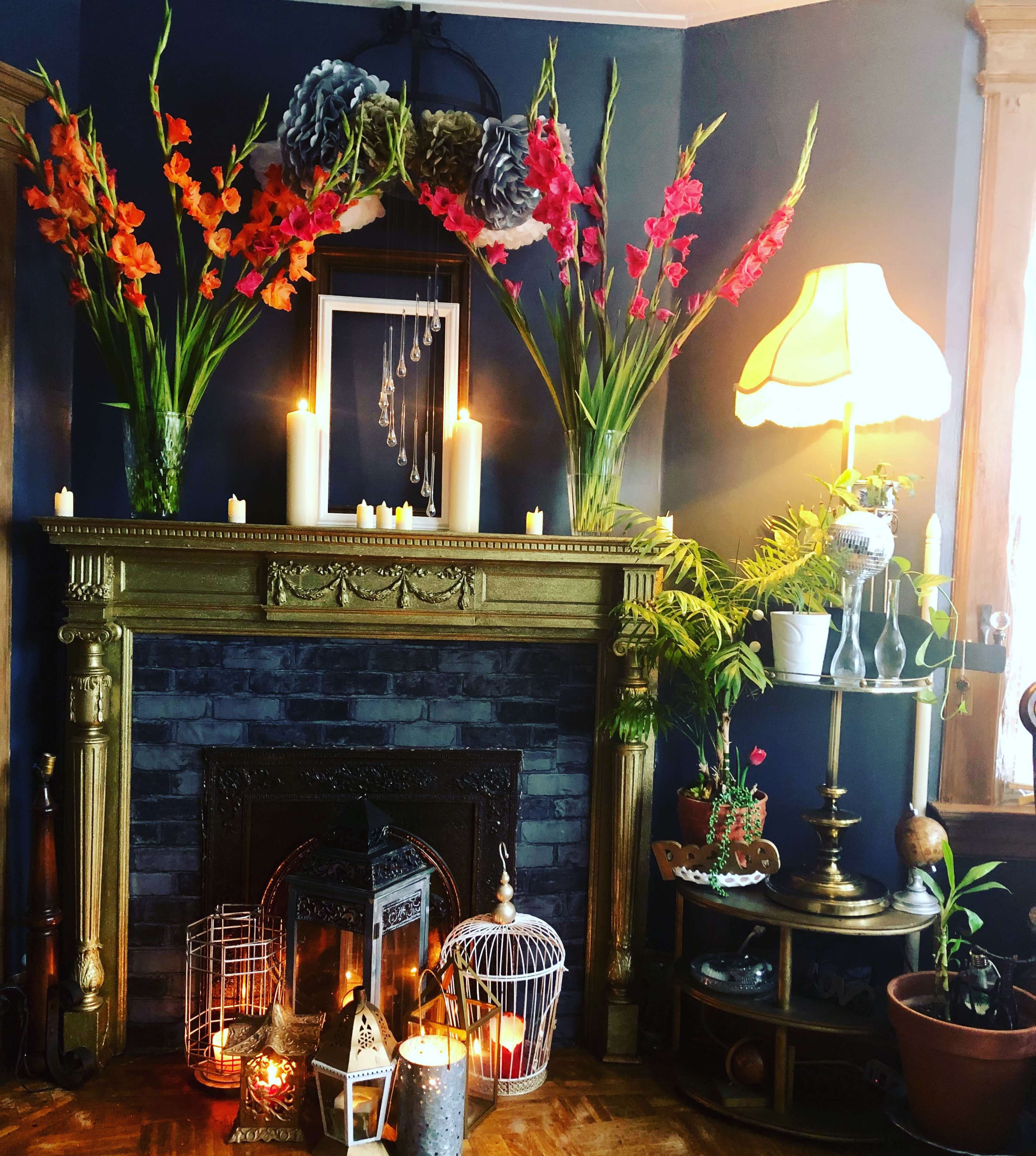 A decorated fireplace with vibrant flowers, candles, and a lamp, surrounded by various plants and ornamental items.
