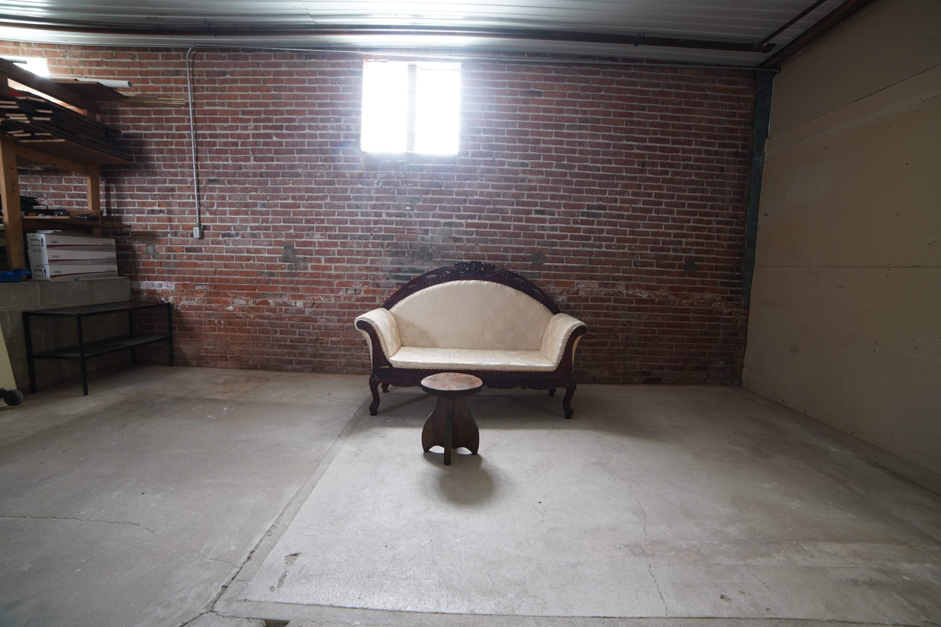 The image shows an empty garage or storage space with a vintage-style sofa and a small table on a concrete floor, against a backdrop of exposed brick walls.