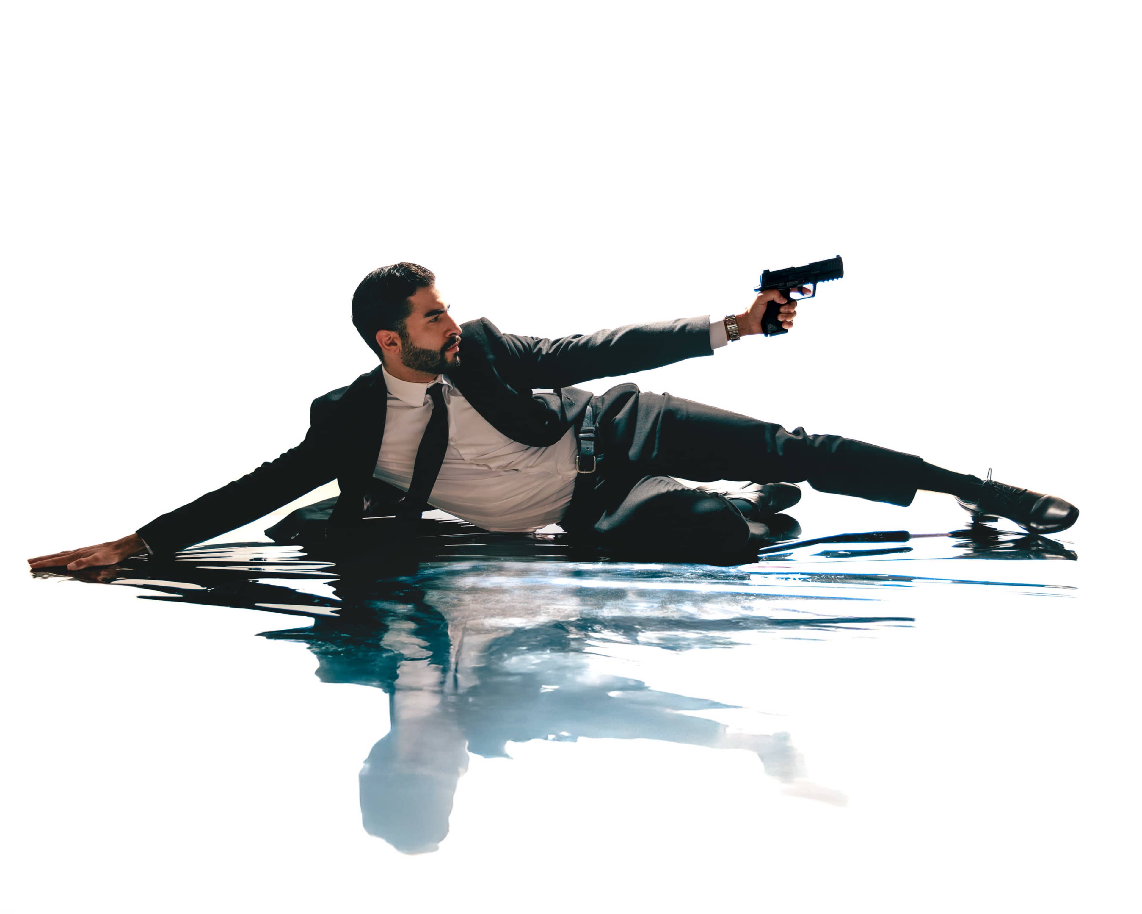 A man in a suit lies on the ground, holding a pistol while reaching out, with a reflective surface beneath him.