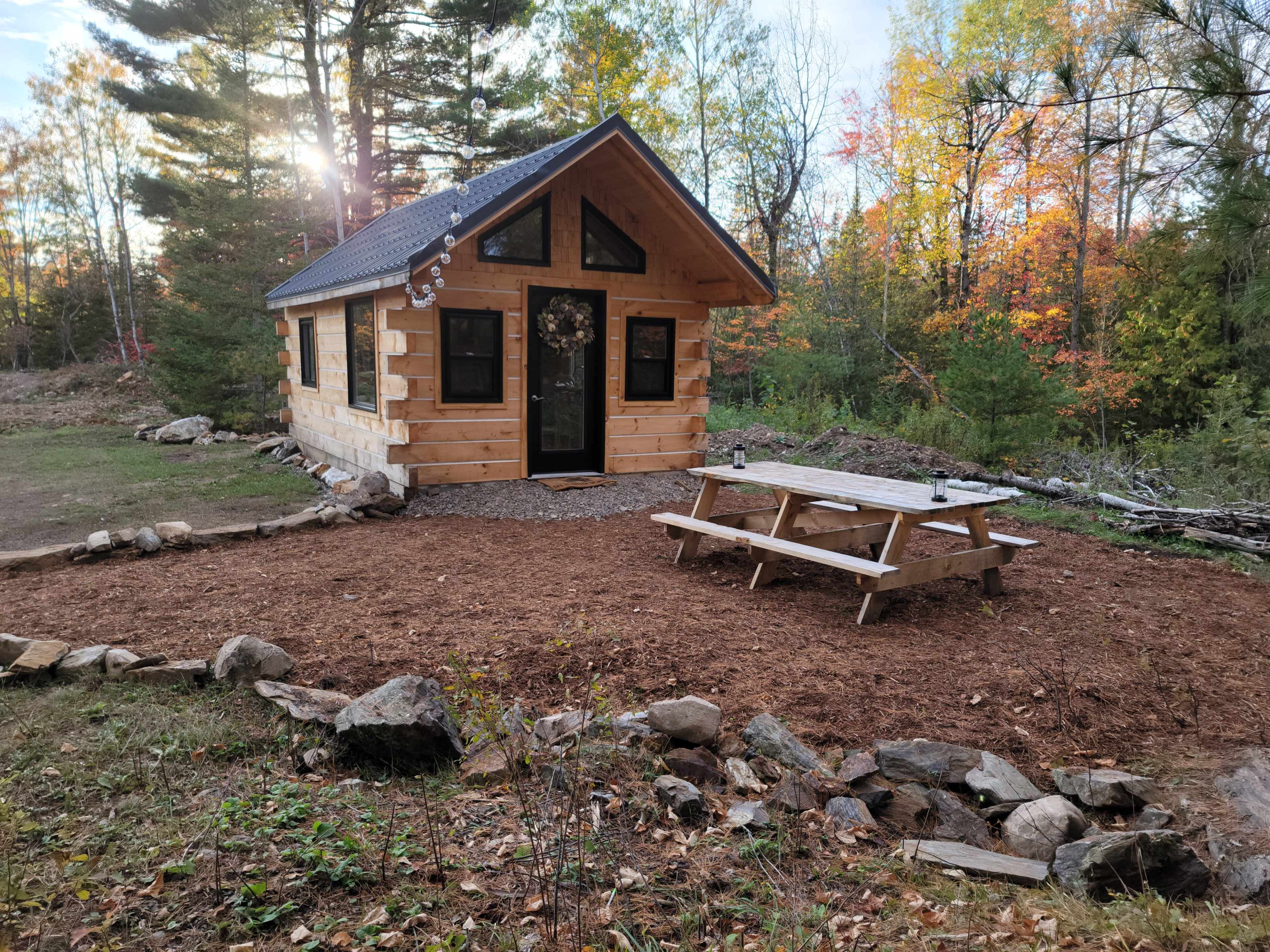 A small, log cabin with a front porch sits in a clearing surrounded by trees, with a picnic table placed nearby on a bed of mulch.