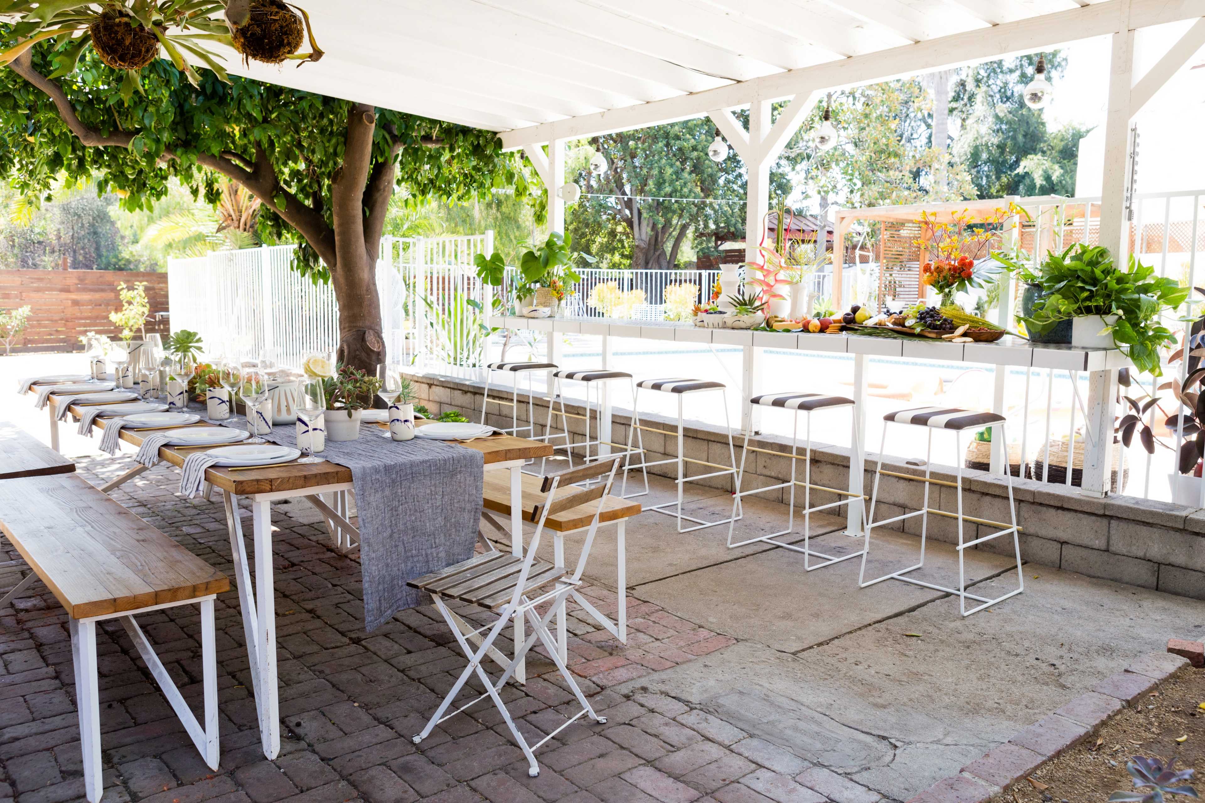 An outdoor dining area features a long table set with tableware and surrounded by greenery, with bar stools positioned at a counter nearby.