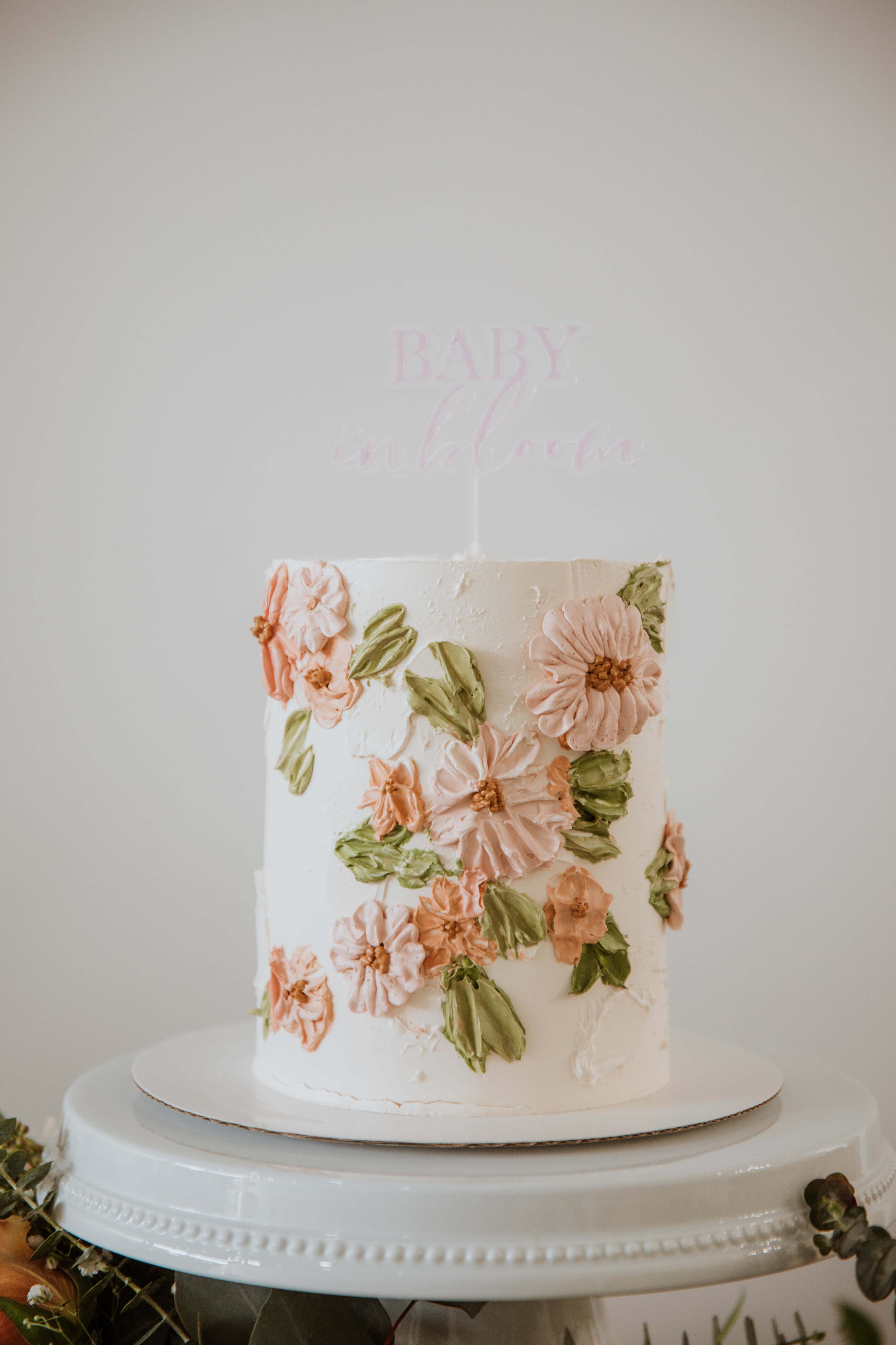 A three-tier white cake is adorned with pink and green floral decorations and features a "Baby" cake topper.