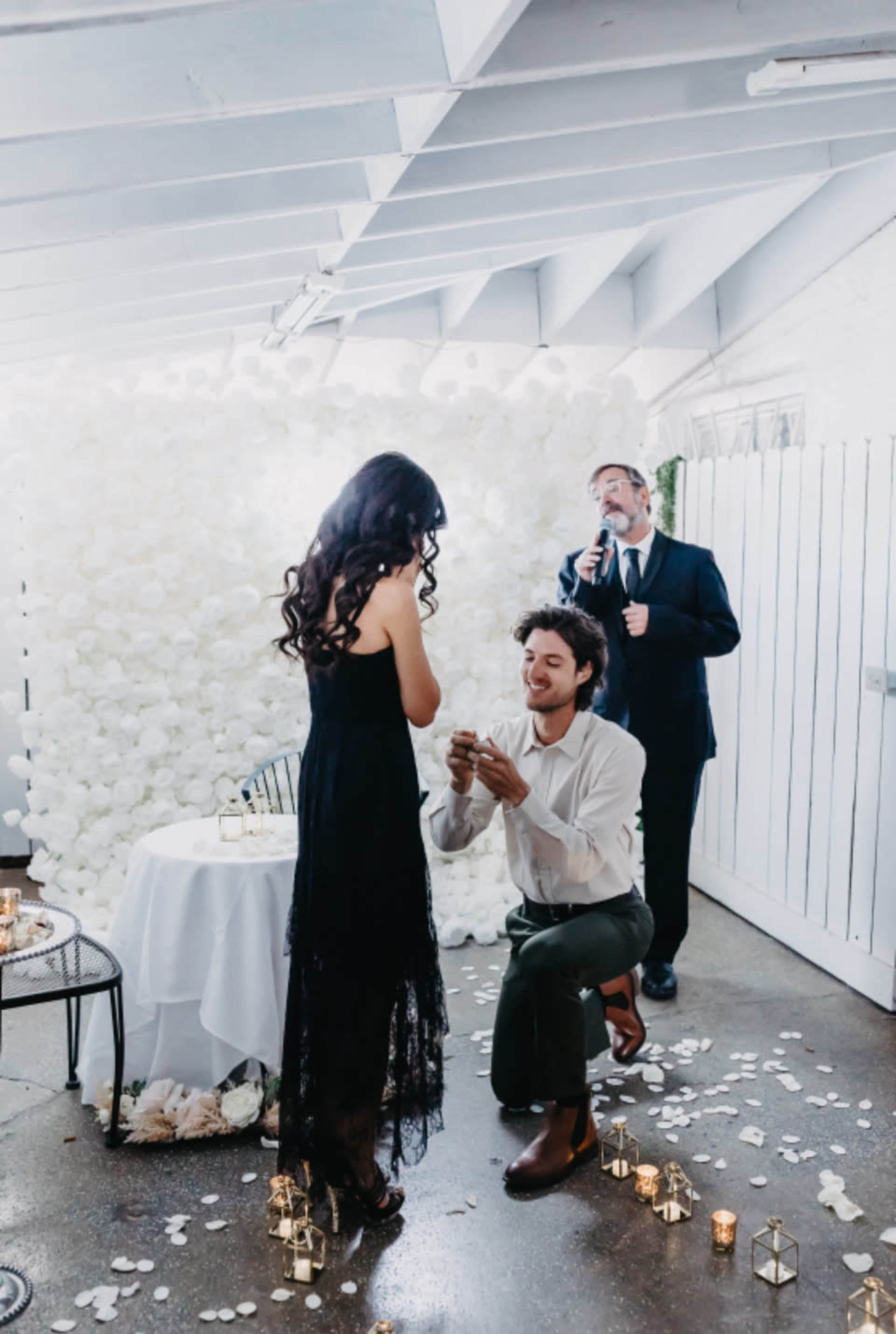 A man is kneeling and proposing to a woman in a decorated indoor space, while another man is singing nearby.