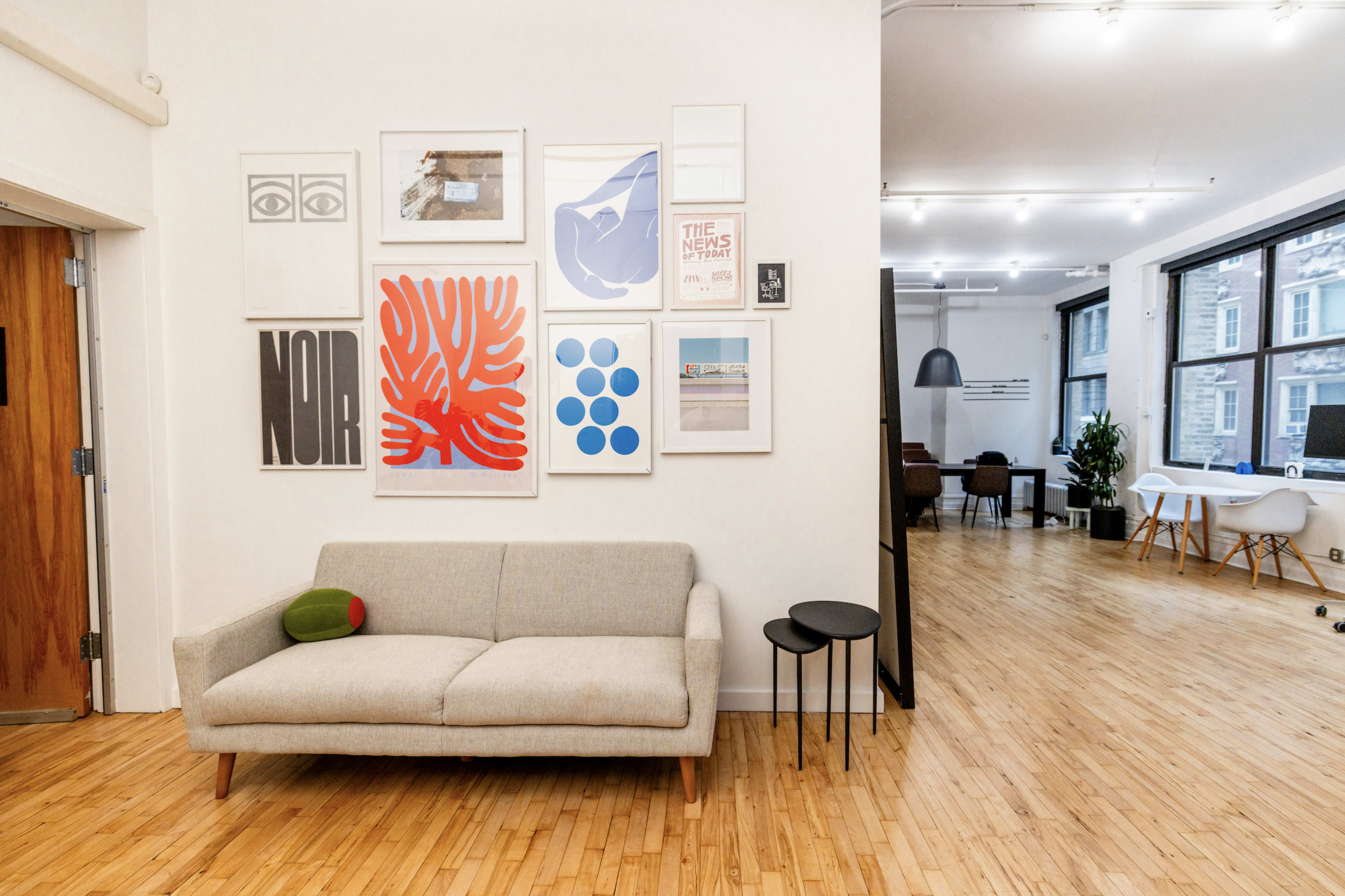 A modern interior space features a sofa against a wall adorned with various framed artworks, while an open area in the background includes tables and chairs.