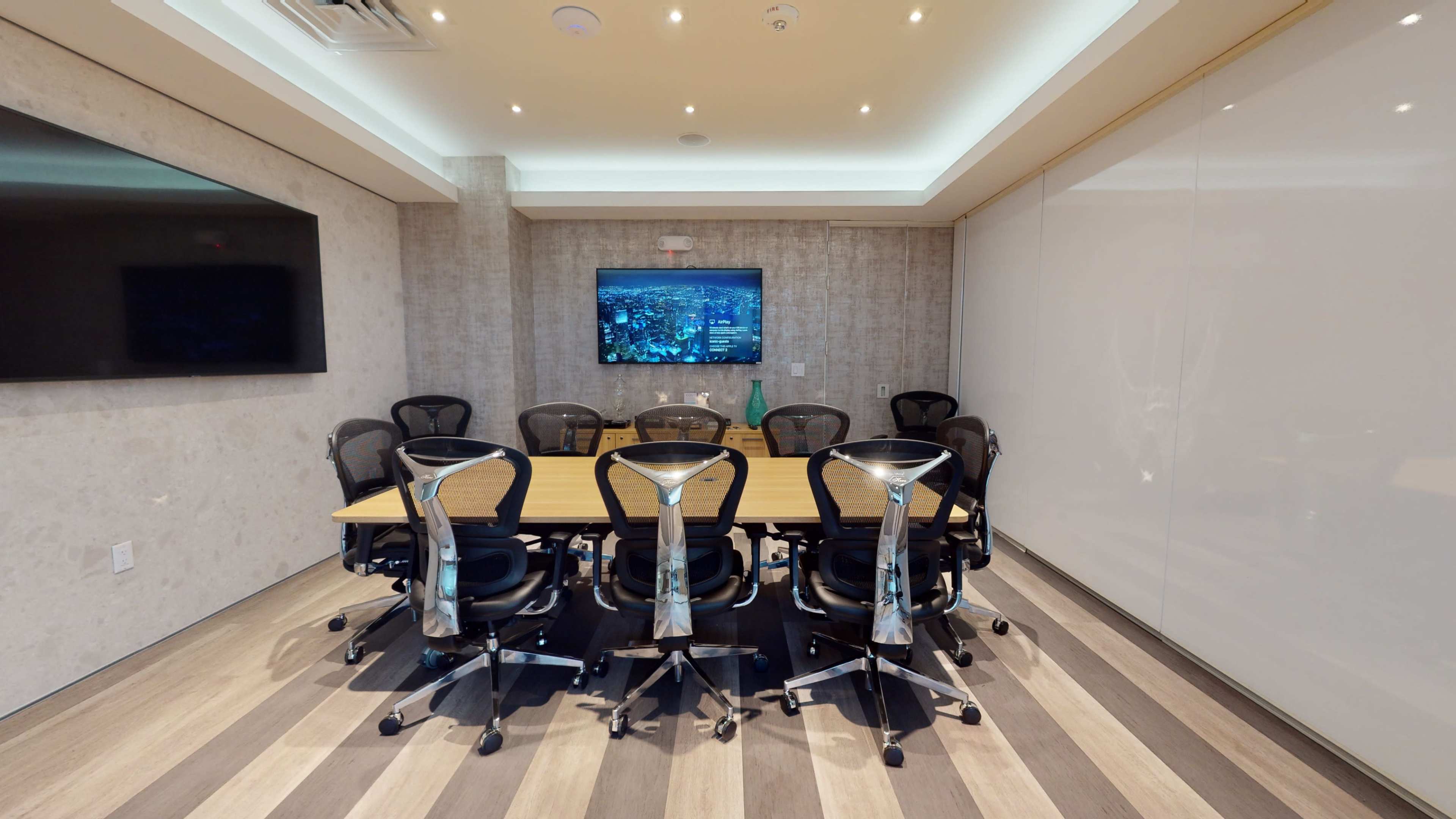A rectangular conference room features a large table surrounded by ergonomic chairs, with a wall-mounted screen displaying visuals.