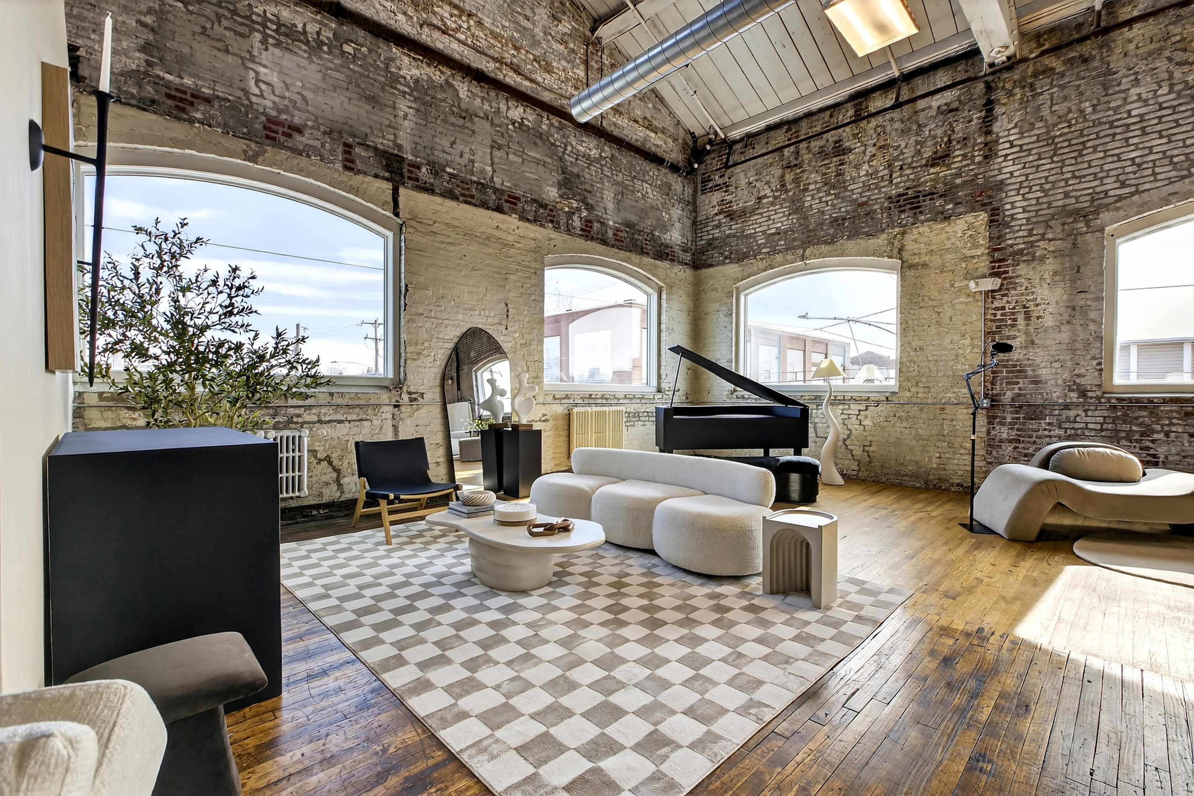 The image shows a spacious, modern living room with large windows, a grand piano, and minimalistic furniture arranged on a checkered rug.