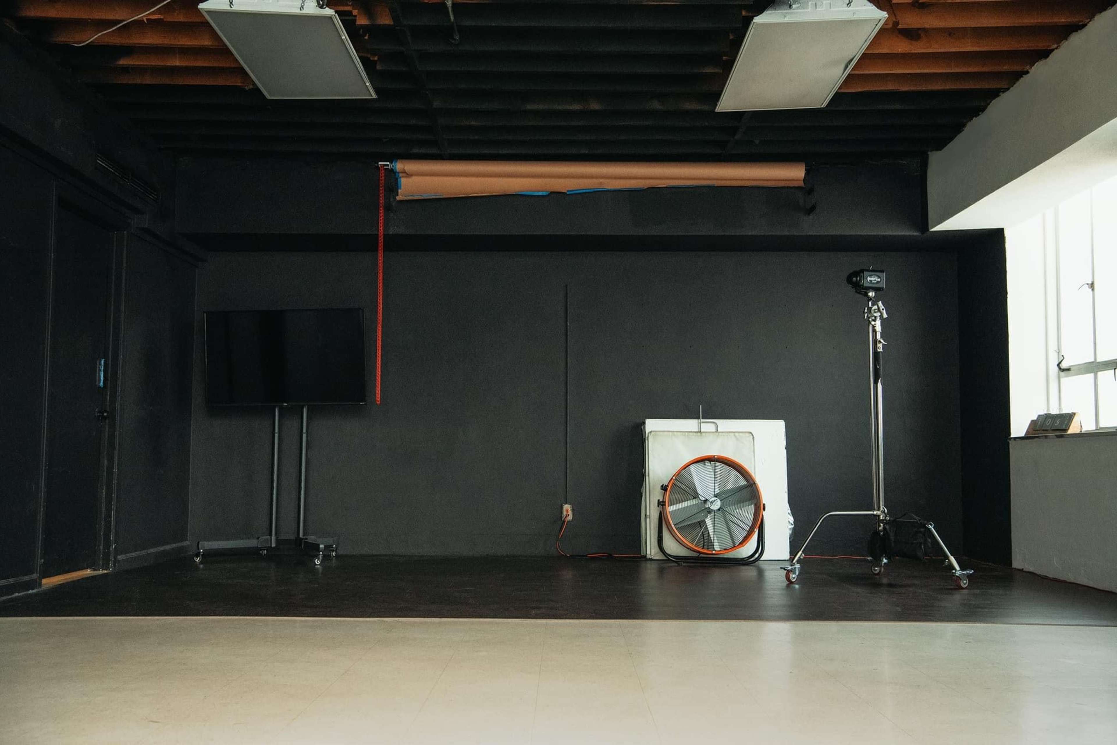 An empty studio space featuring a mounted television, a large industrial fan, and a lighting stand against a dark wall.
