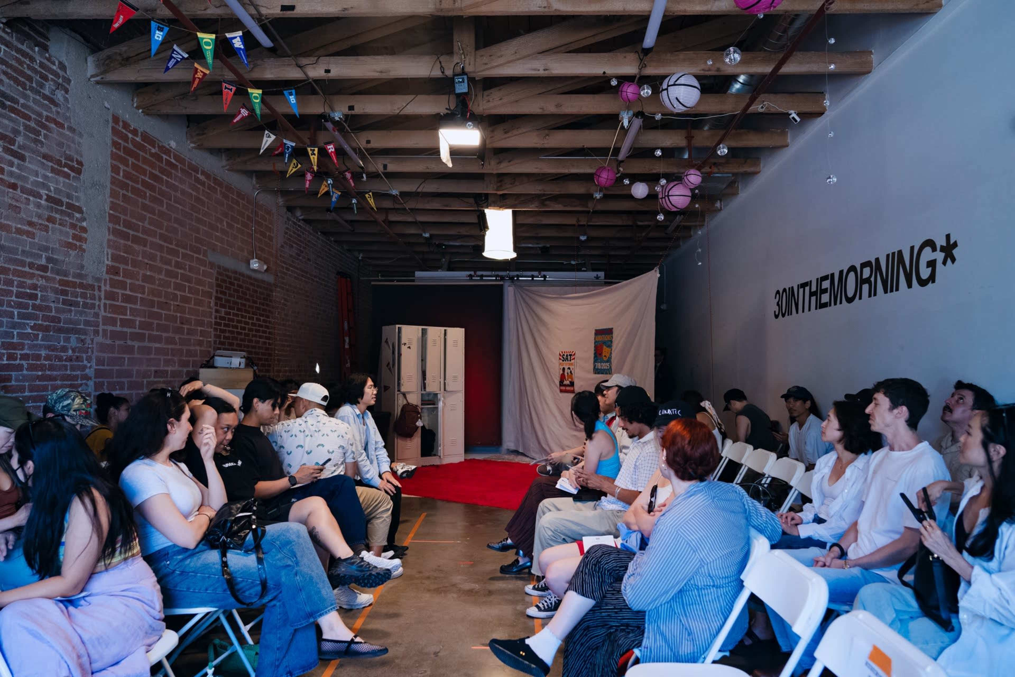 A group of people sits in white chairs arranged in rows inside a spacious, industrial-style room adorned with colorful decorations and lockers at the back.