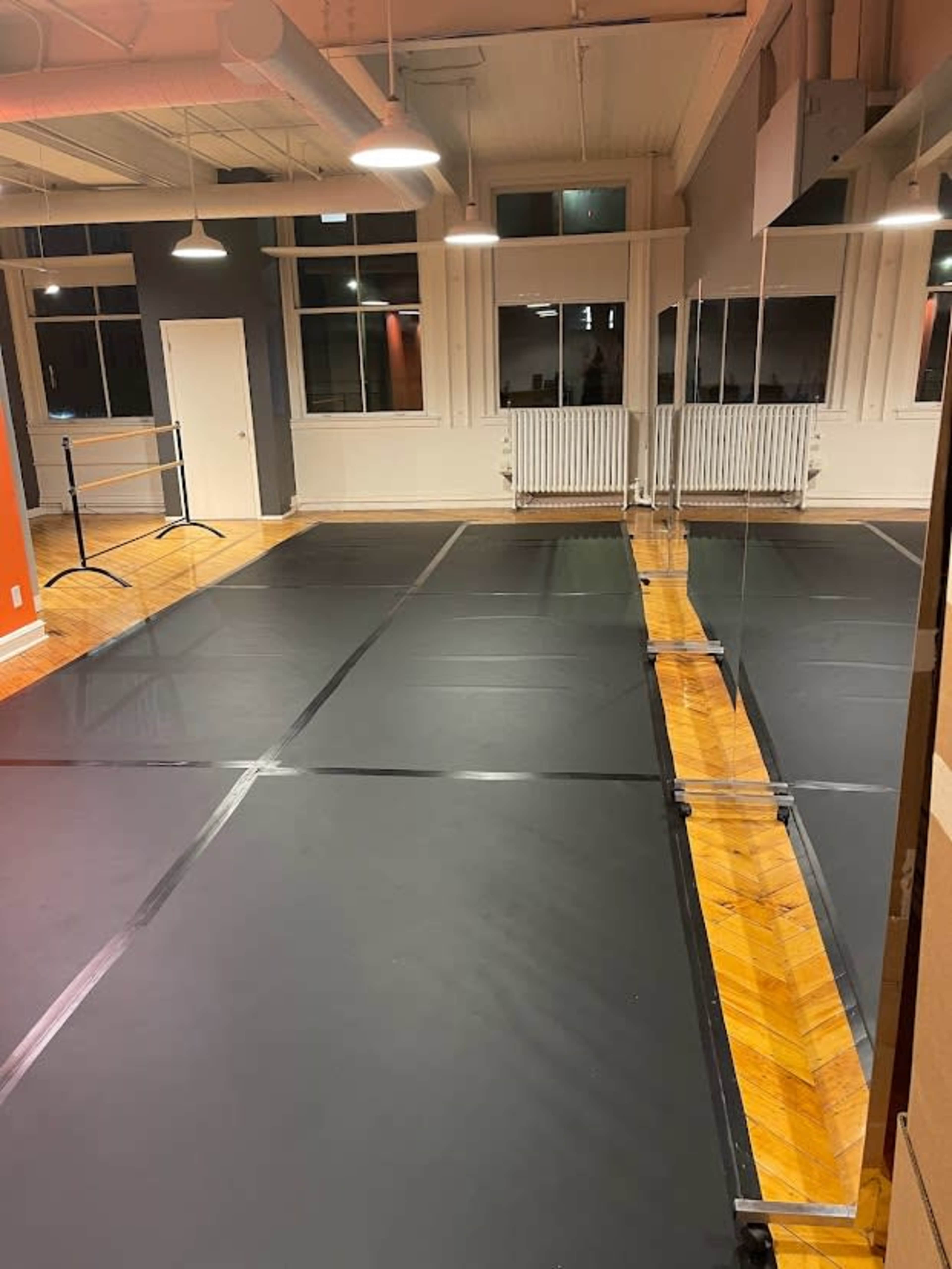 A dance studio with a wooden floor, black mats laid out, mirrors on one wall, and large windows allowing light in.