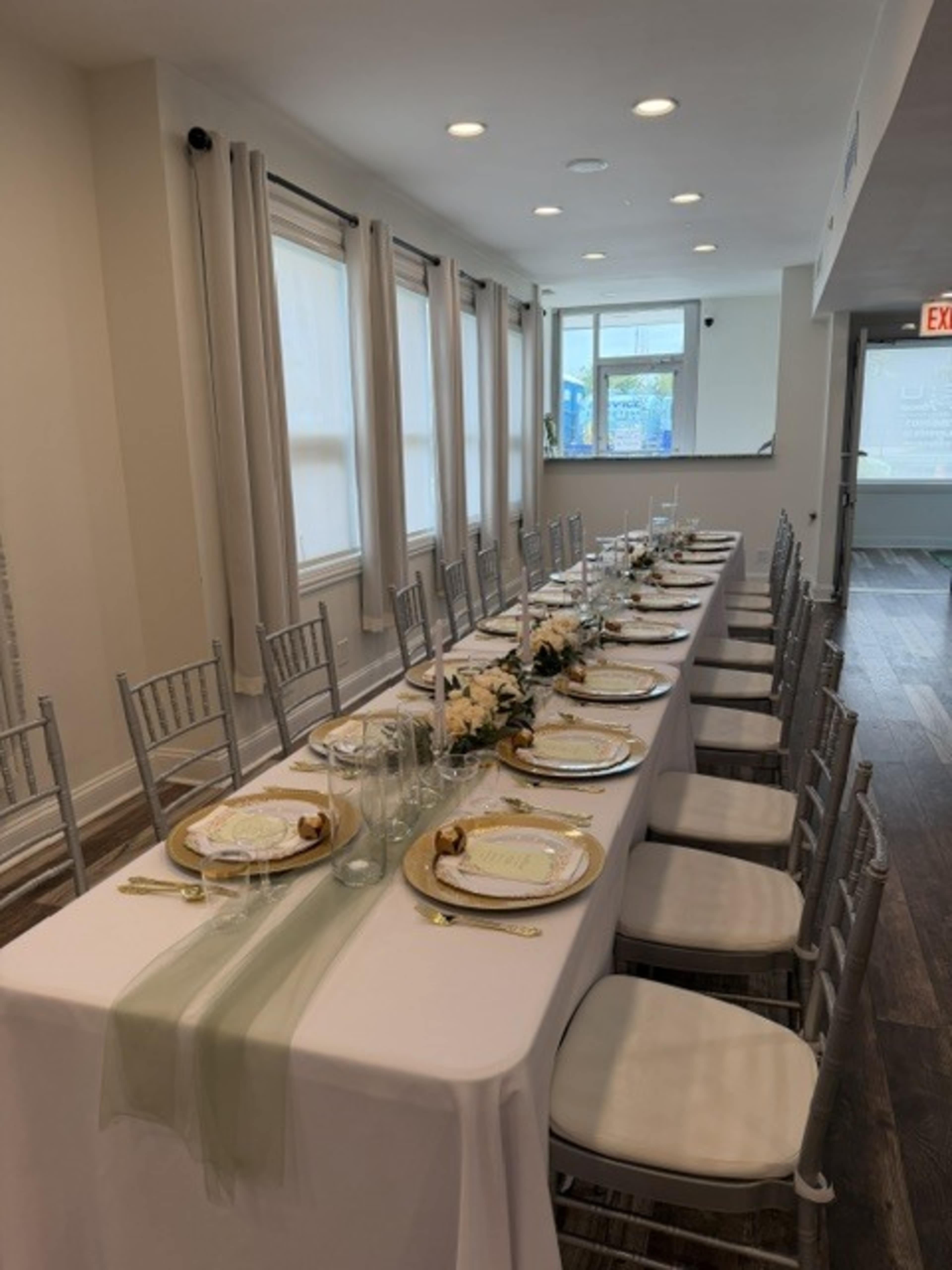 A long dining table is elegantly set with plates and glassware, surrounded by silver chairs in a bright, spacious room with large windows.