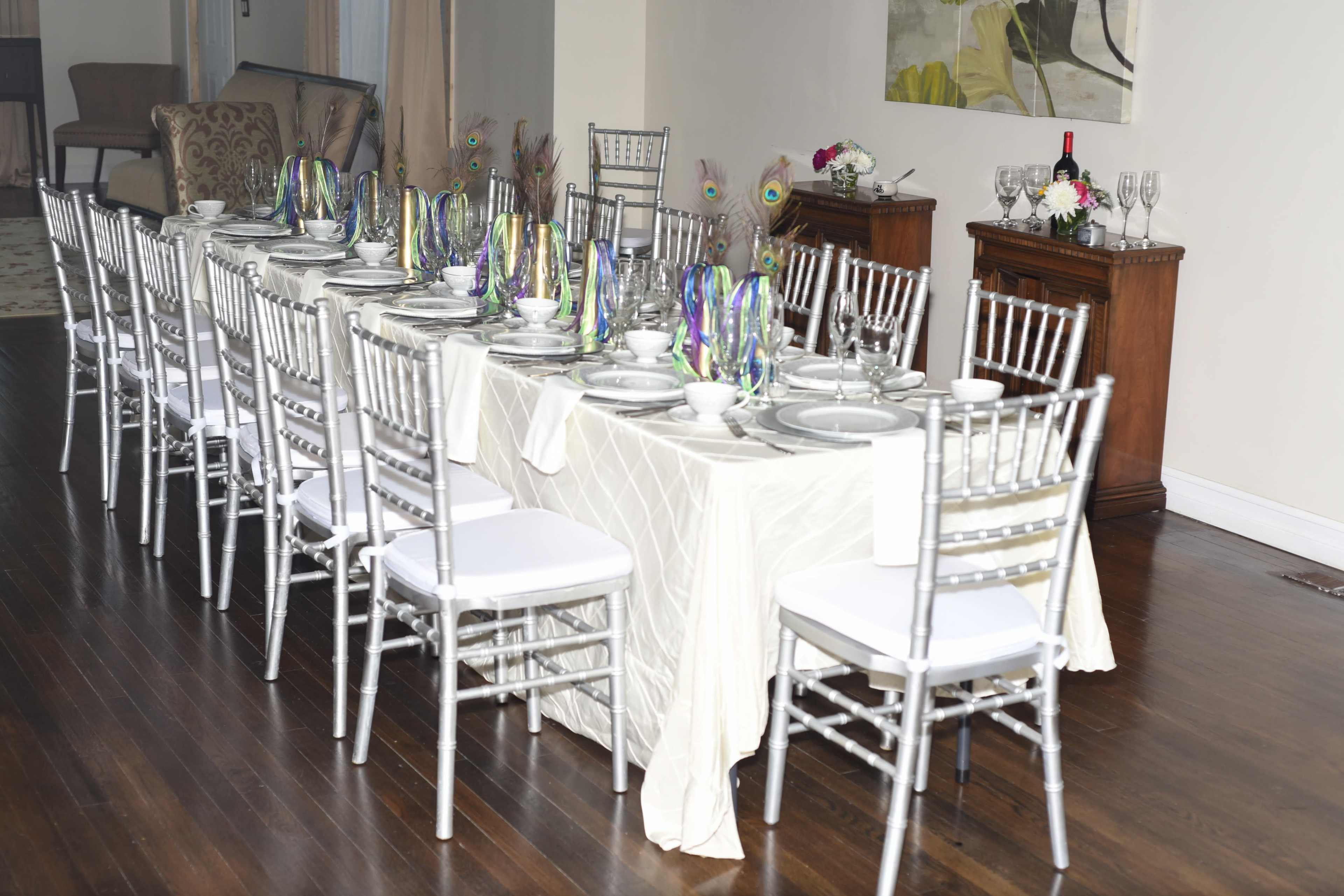 A long dining table is set with fine china and decorated with peacock feather centerpieces, surrounded by silver chiavari chairs.
