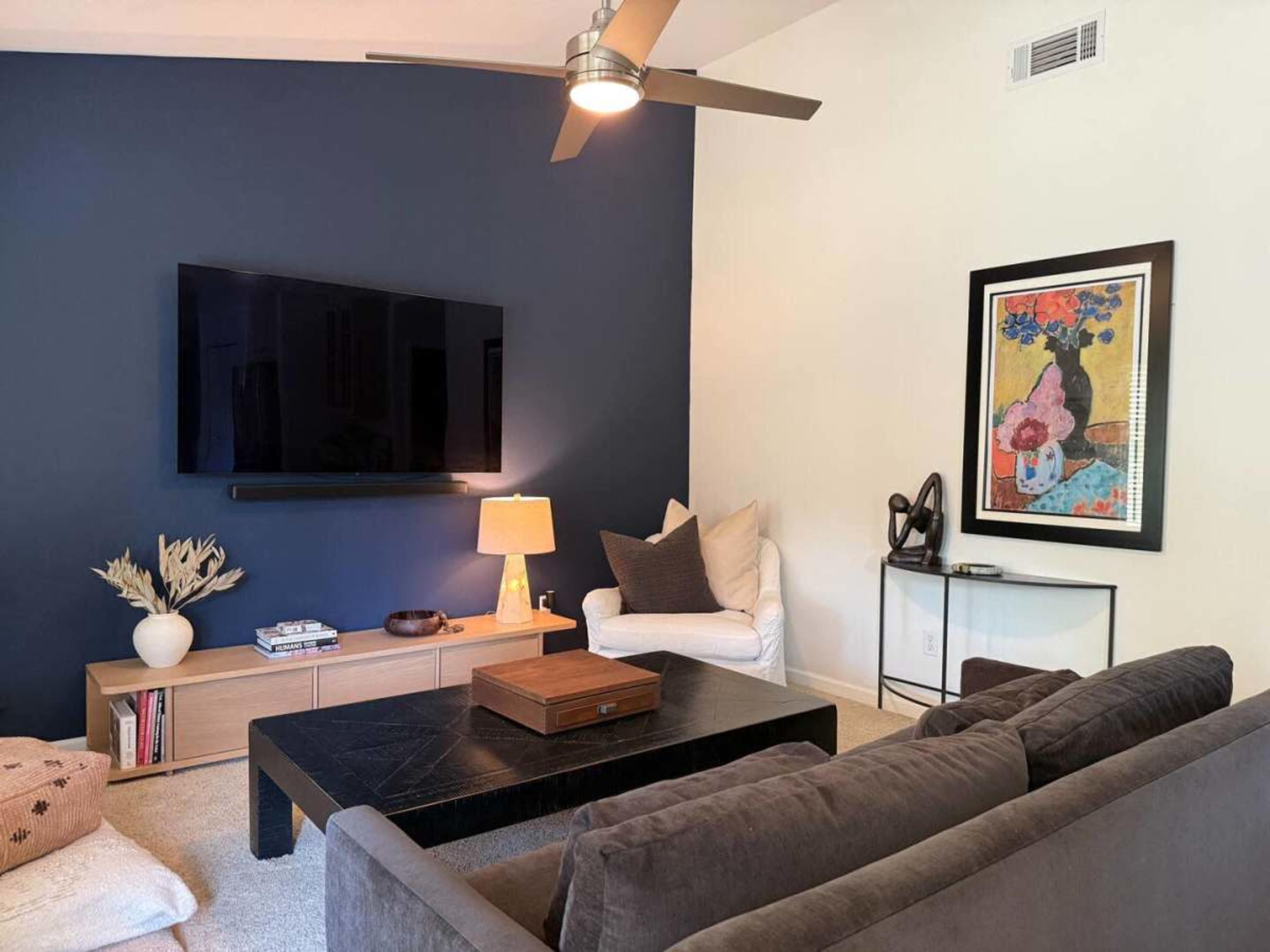 The image shows a modern living room with a dark blue accent wall, a large flat-screen TV mounted above a light wooden media console, a white armchair, and a gray sofa facing a black coffee table.