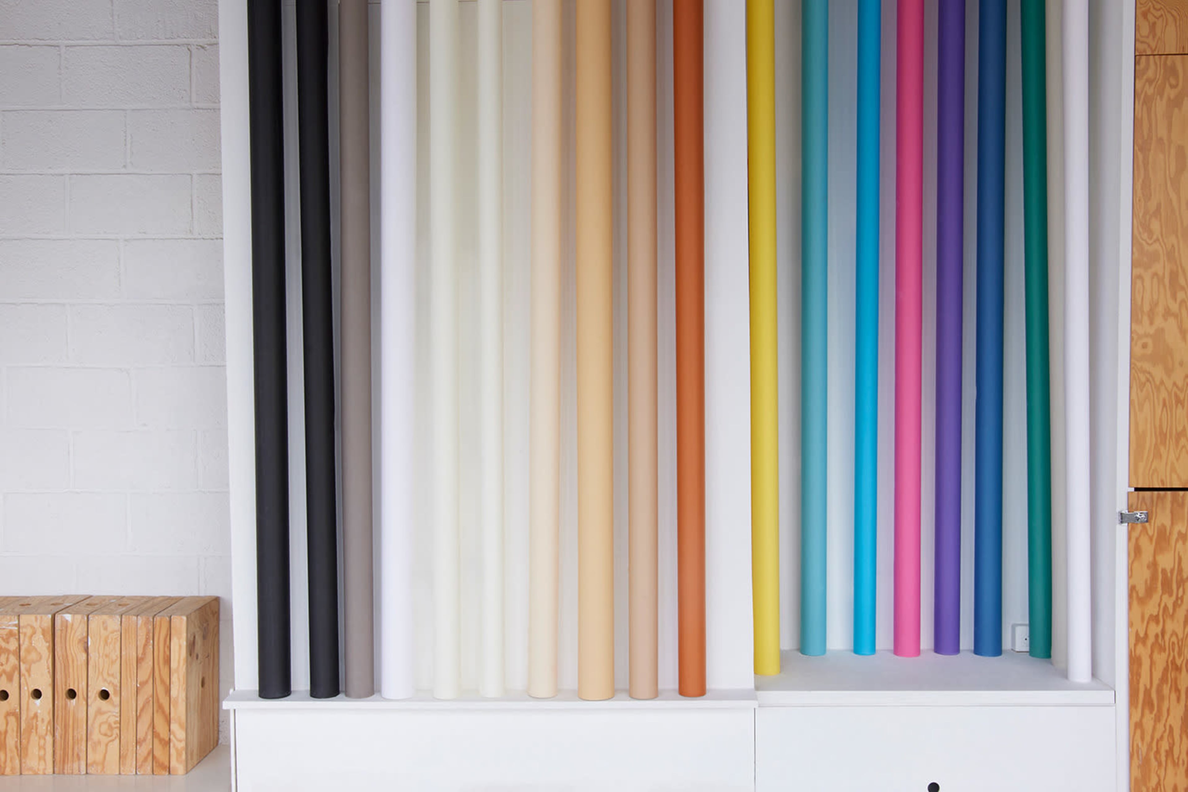 The image features a wall displaying a variety of vertically arranged rolls of colored paper in several hues.