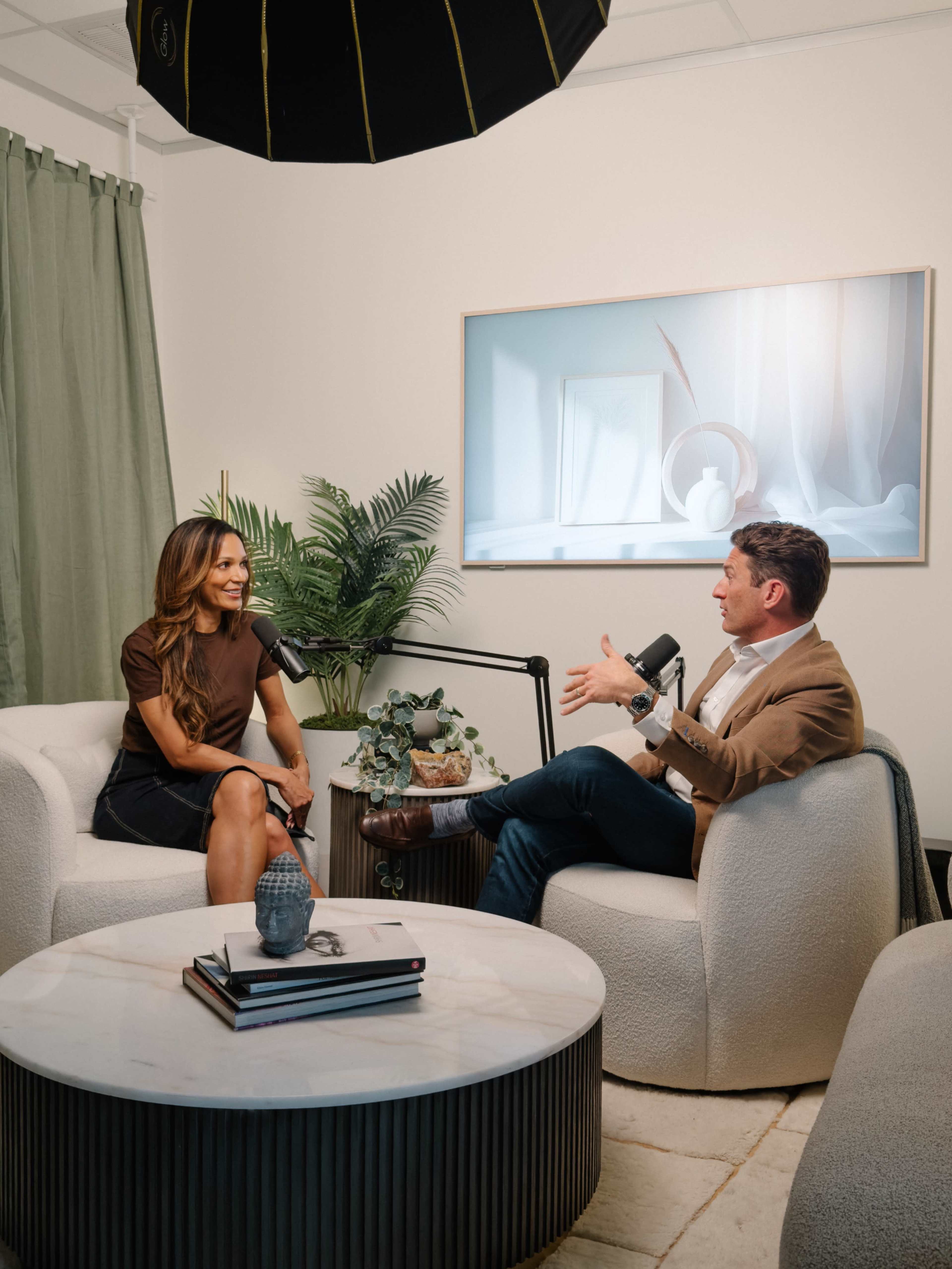 A man and a woman are seated in a modern conversation space, engaging with each other while decorative plants and a photographic artwork are in the background.