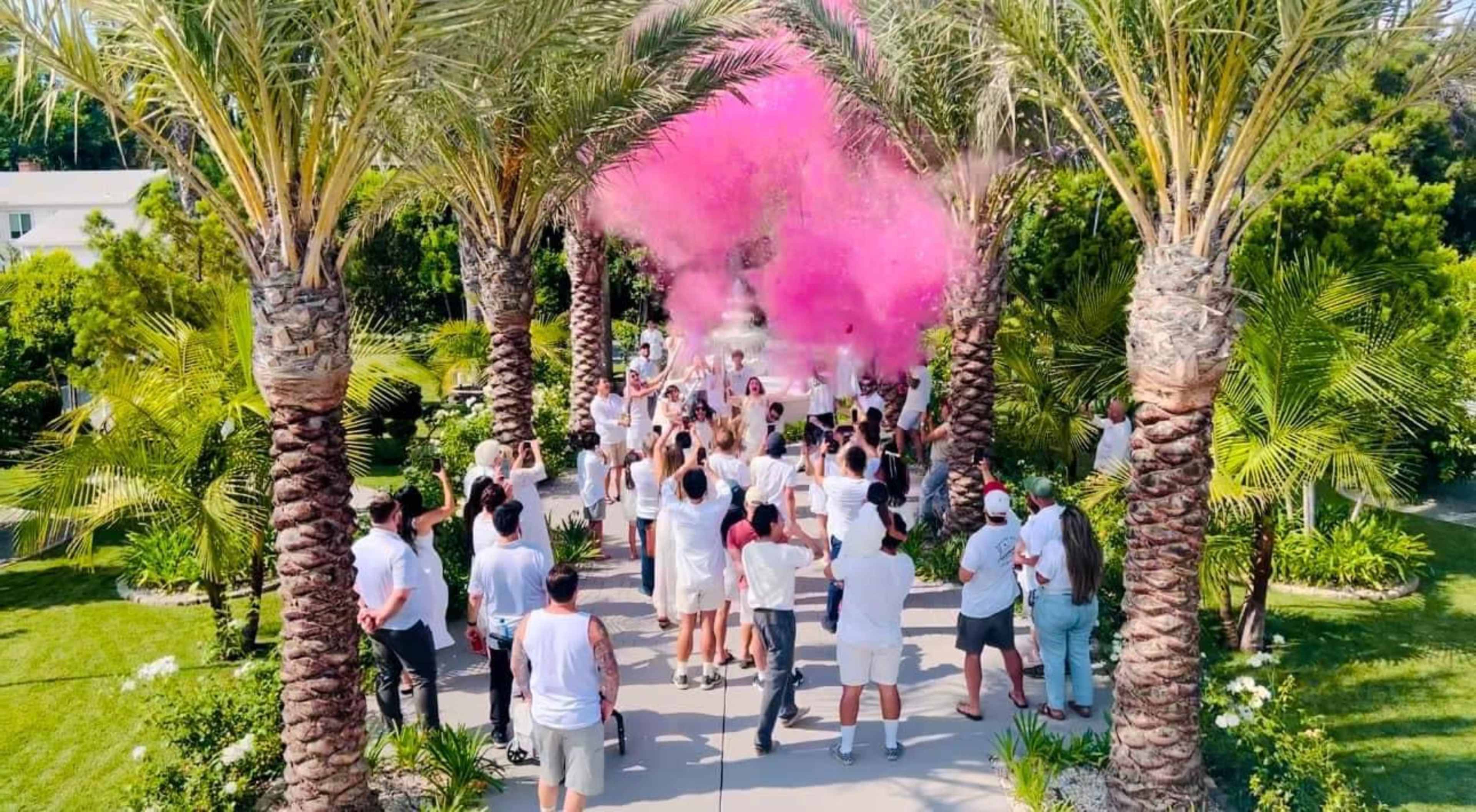 A large group of people wearing white clothing gathers under palm trees as pink smoke fills the air, creating a vibrant scene in a garden setting.