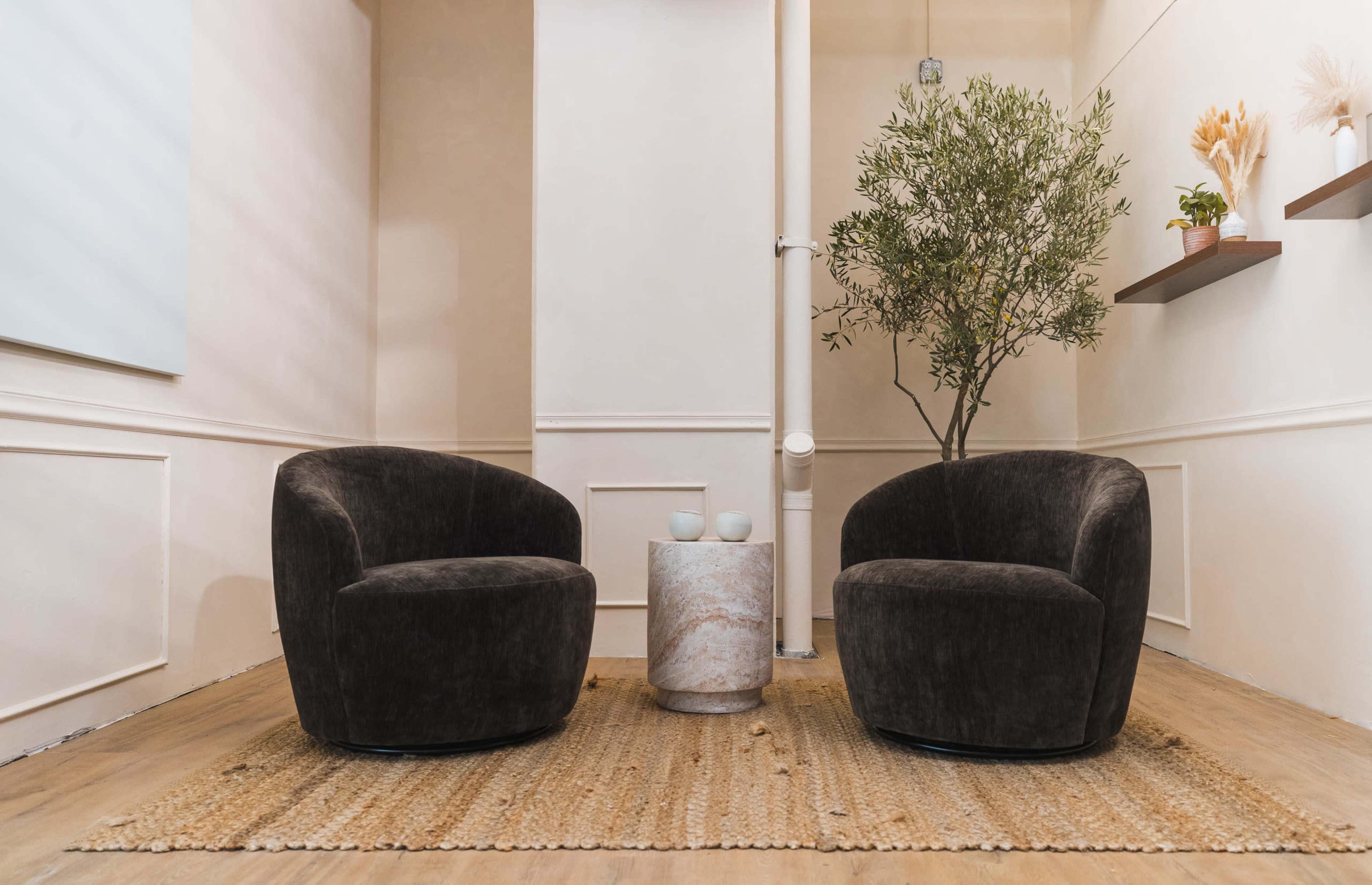 The image shows two curved, dark chairs flanking a round marble table on a woven rug, with a decorative plant in the background and minimalistic wall decor.