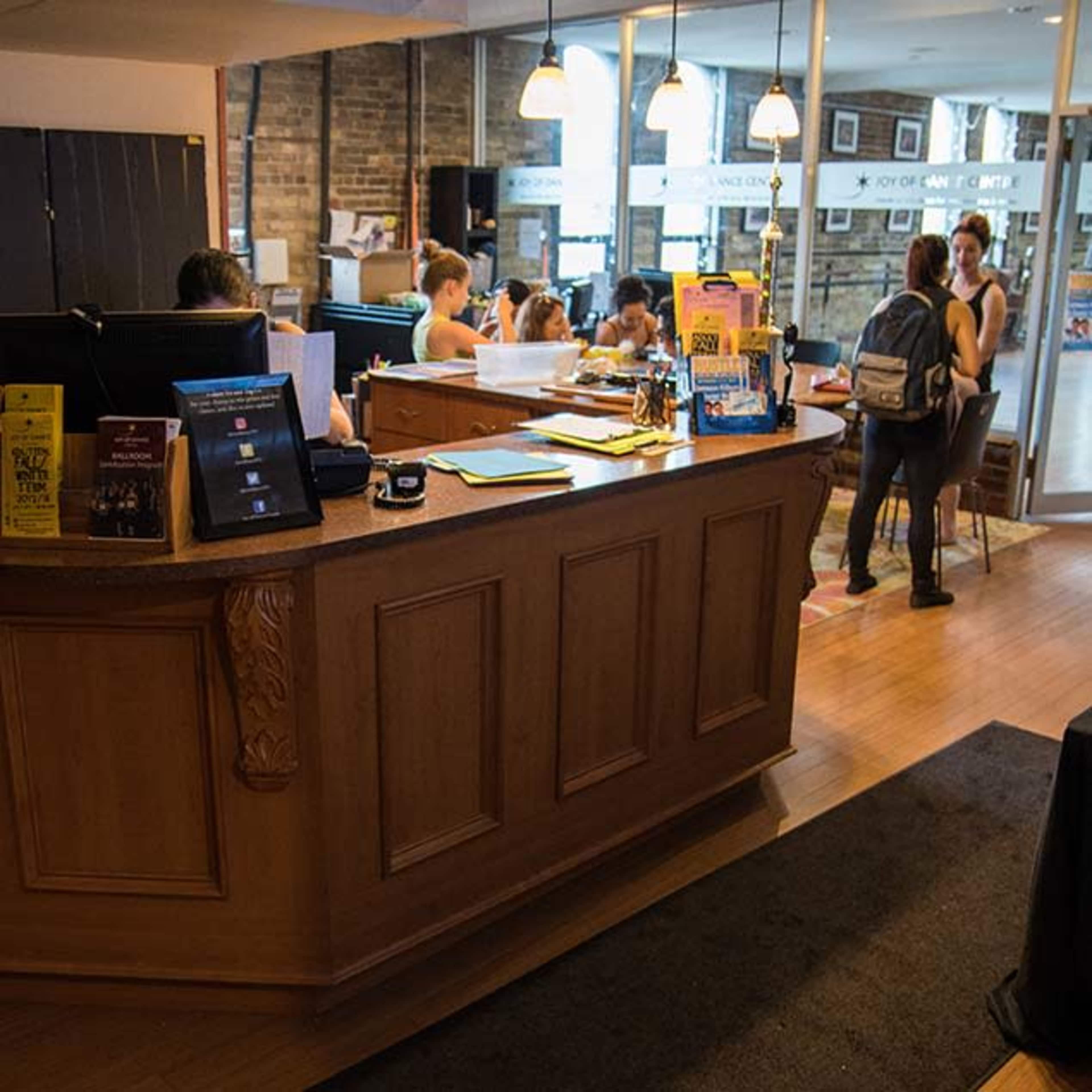 A wooden reception desk stands in the foreground of a brightly lit office space, with several people engaged in various activities in the background.