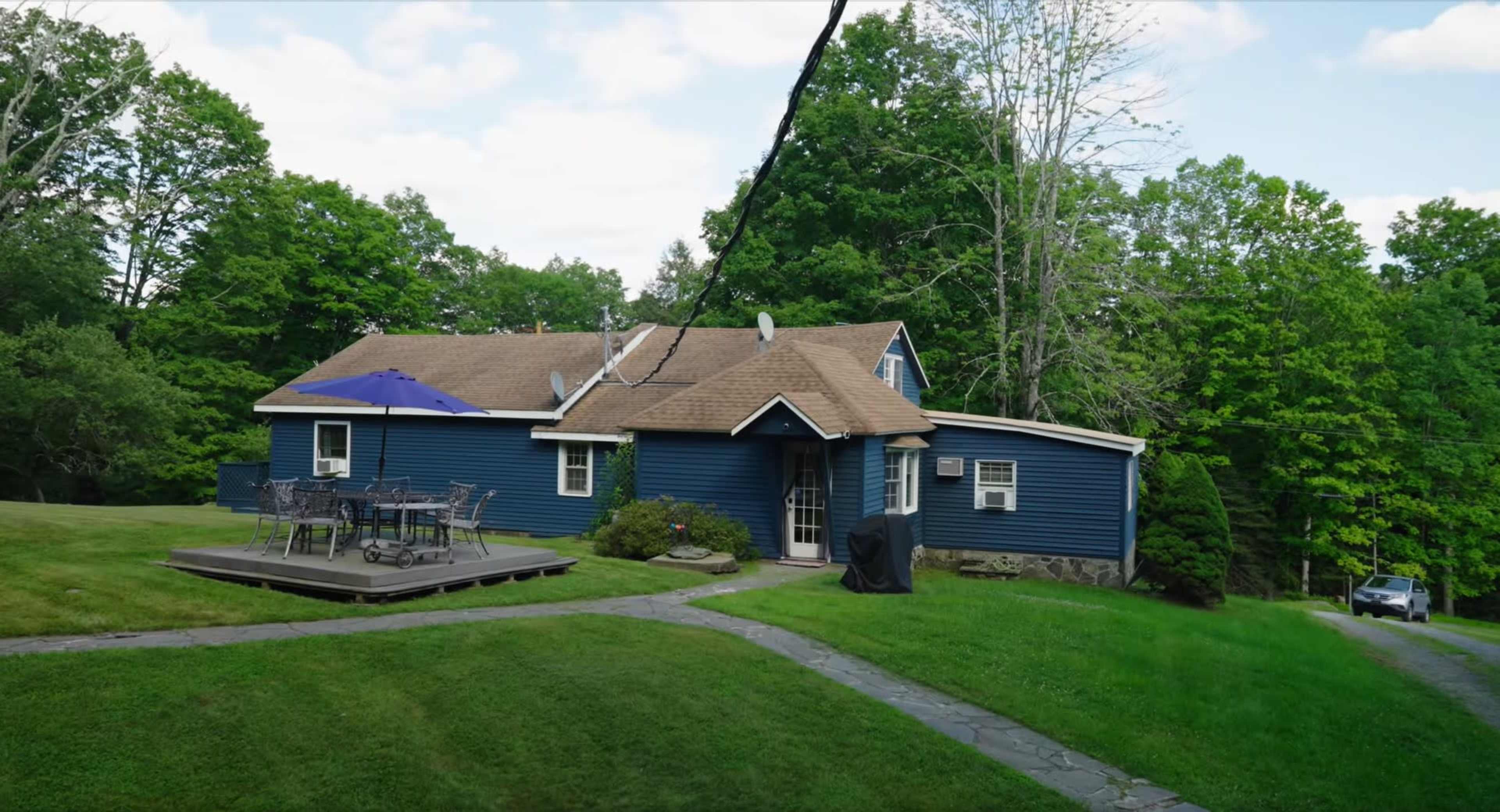 A blue house with a brown roof, surrounded by green trees and grass, features a deck and outdoor seating area.