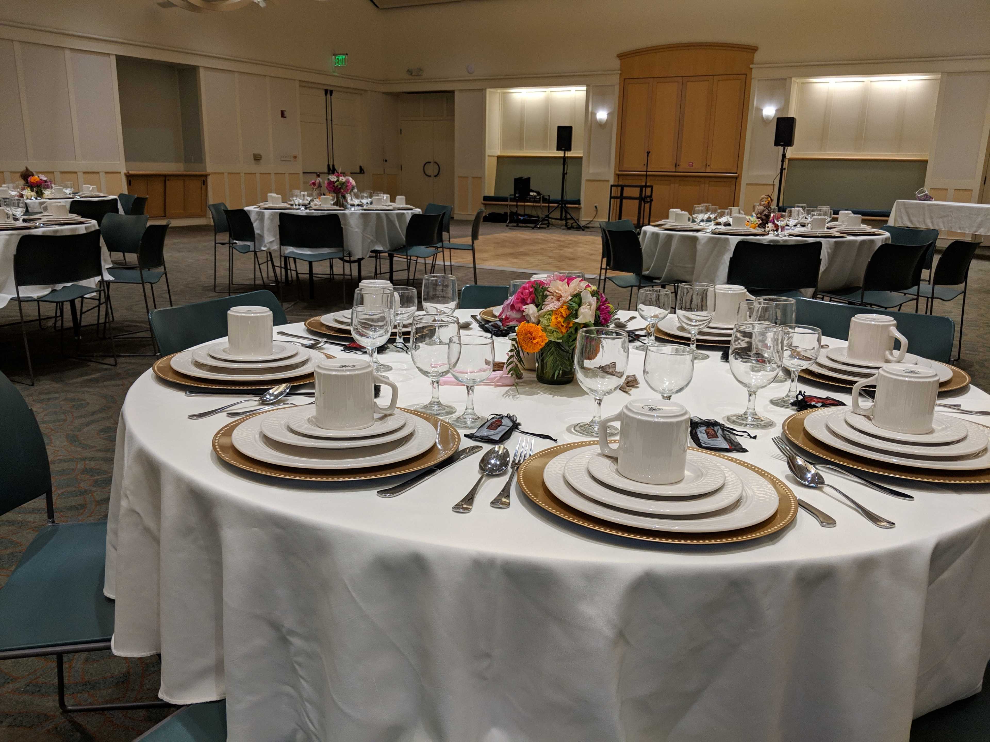 A dining room set for a formal event features round tables with white tablecloths, gold chargers, and dishes arranged neatly, surrounded by green chairs.