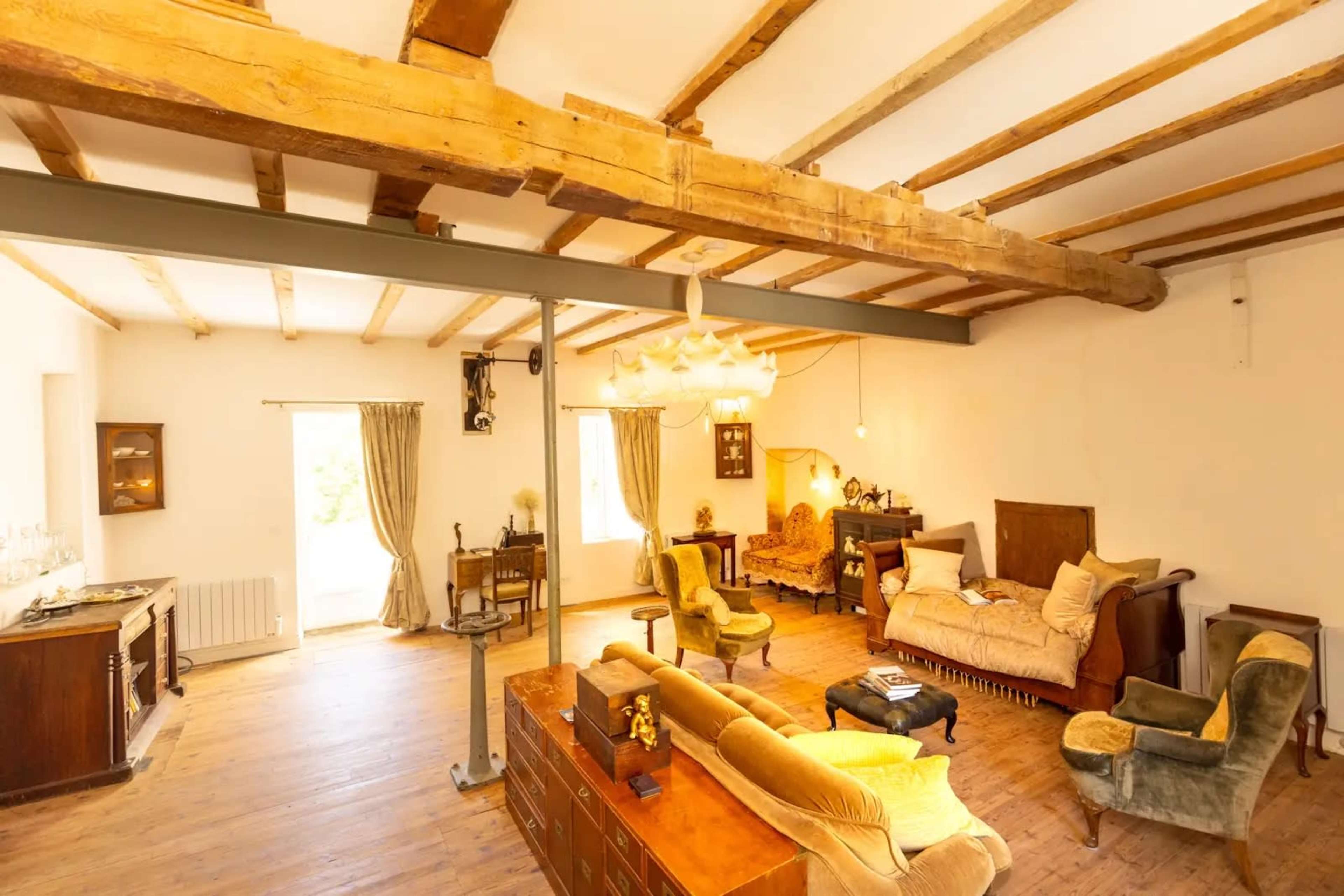 The image shows a spacious living room with wooden beams, light-colored walls, and a mix of vintage furniture arranged around a central coffee table.