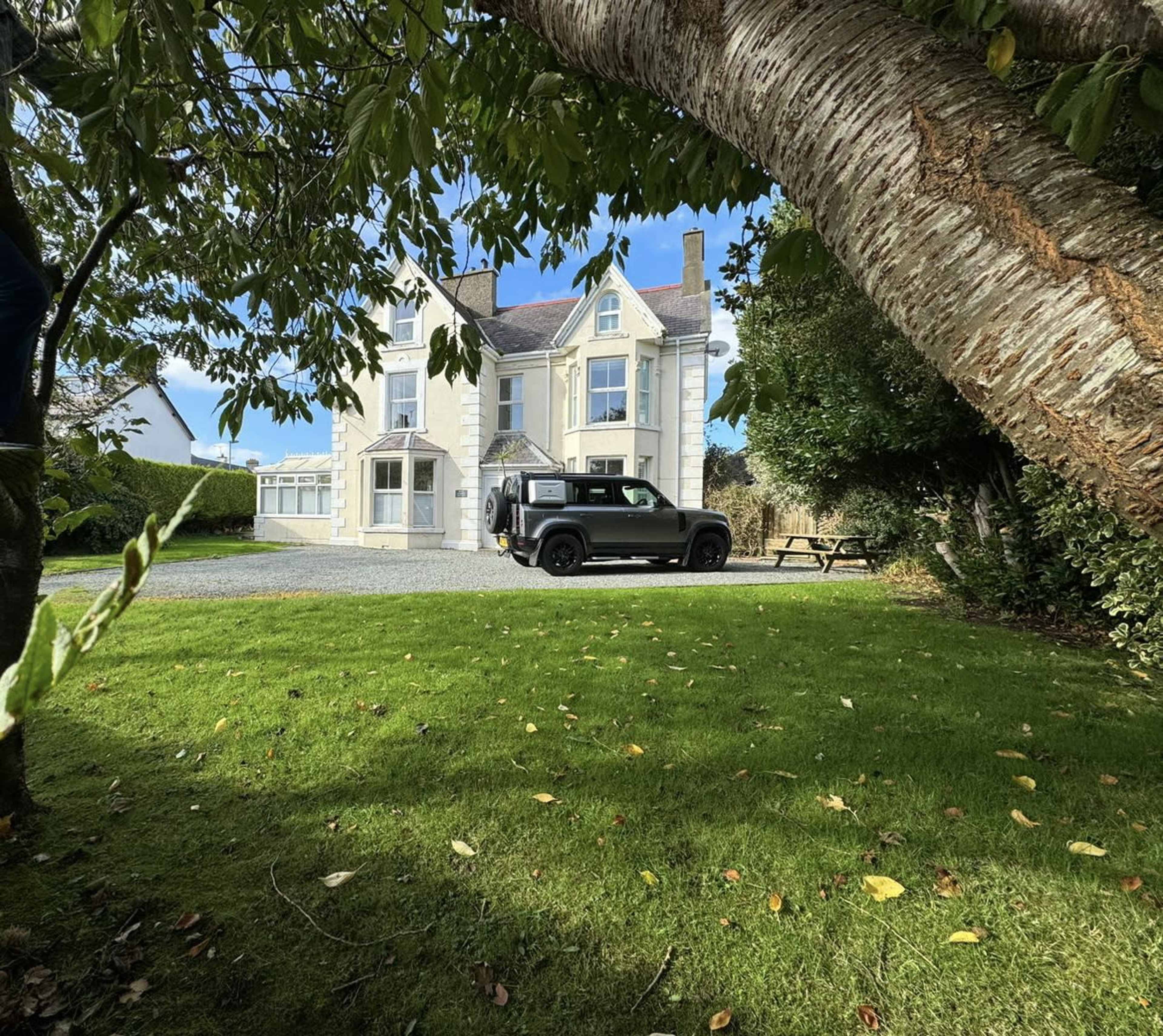 A black vehicle is parked in front of a large, traditional-style house surrounded by a well-kept lawn and trees.