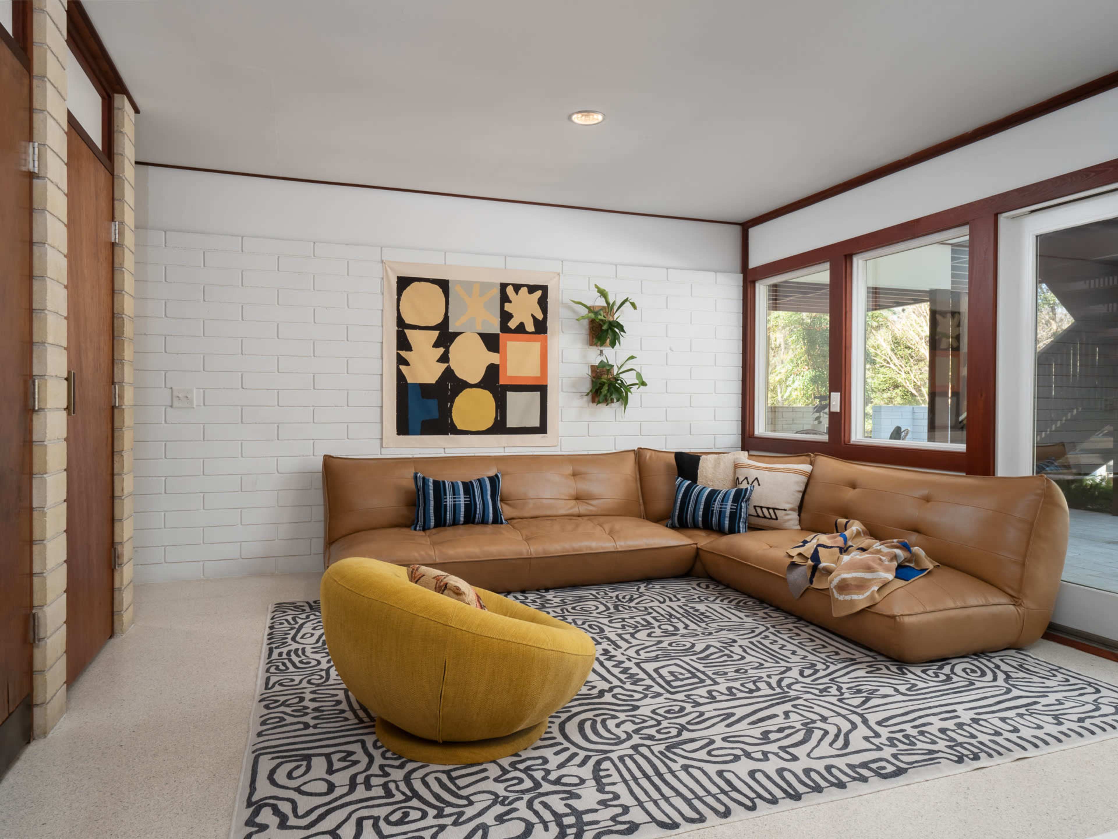A modern living room features a curved brown leather sectional sofa, a patterned area rug, a yellow accent chair, and colorful wall art above a plant.