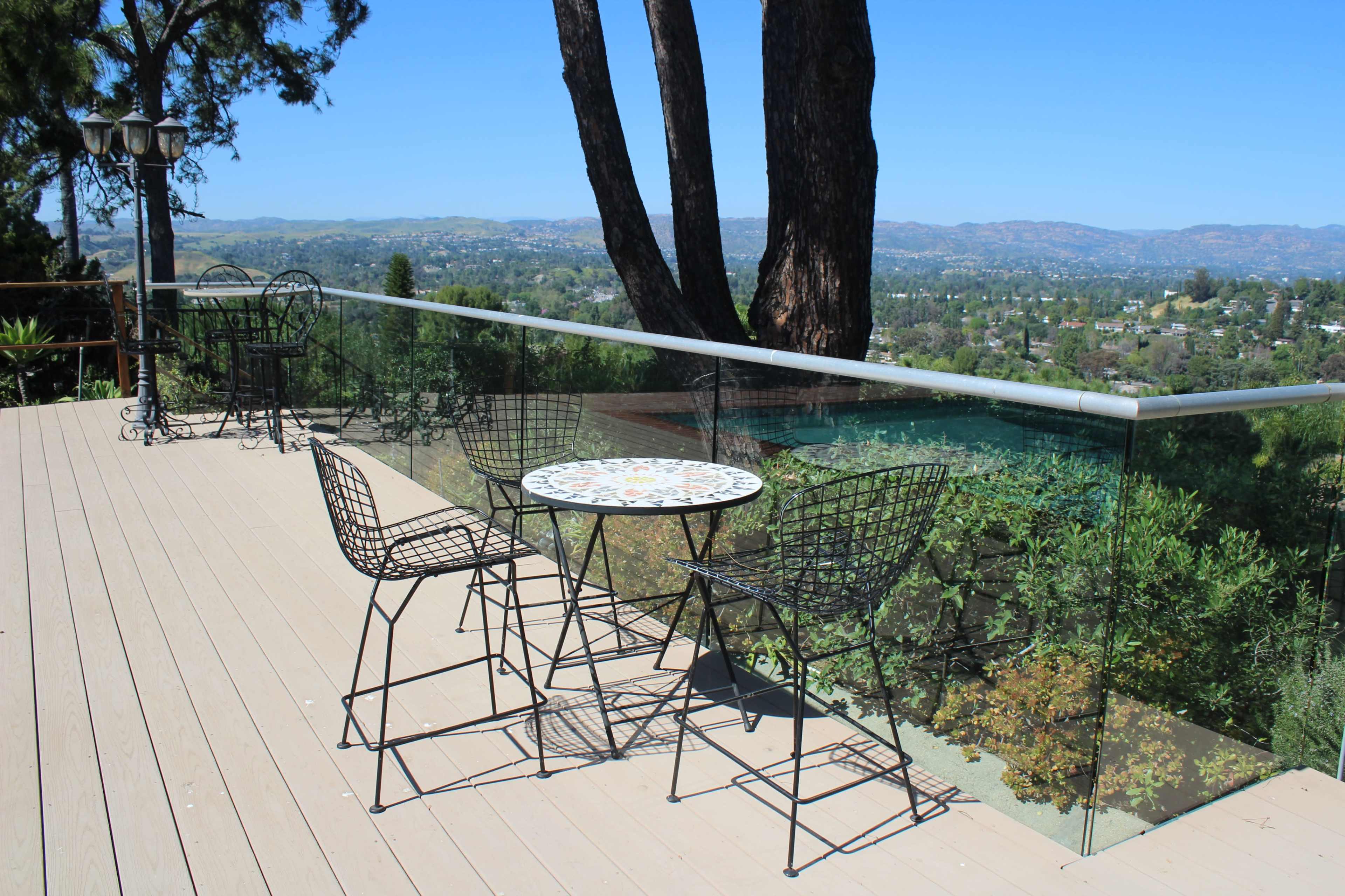 A patio with a round table and two chairs overlooks a scenic valley and distant mountains, framed by tall trees.
