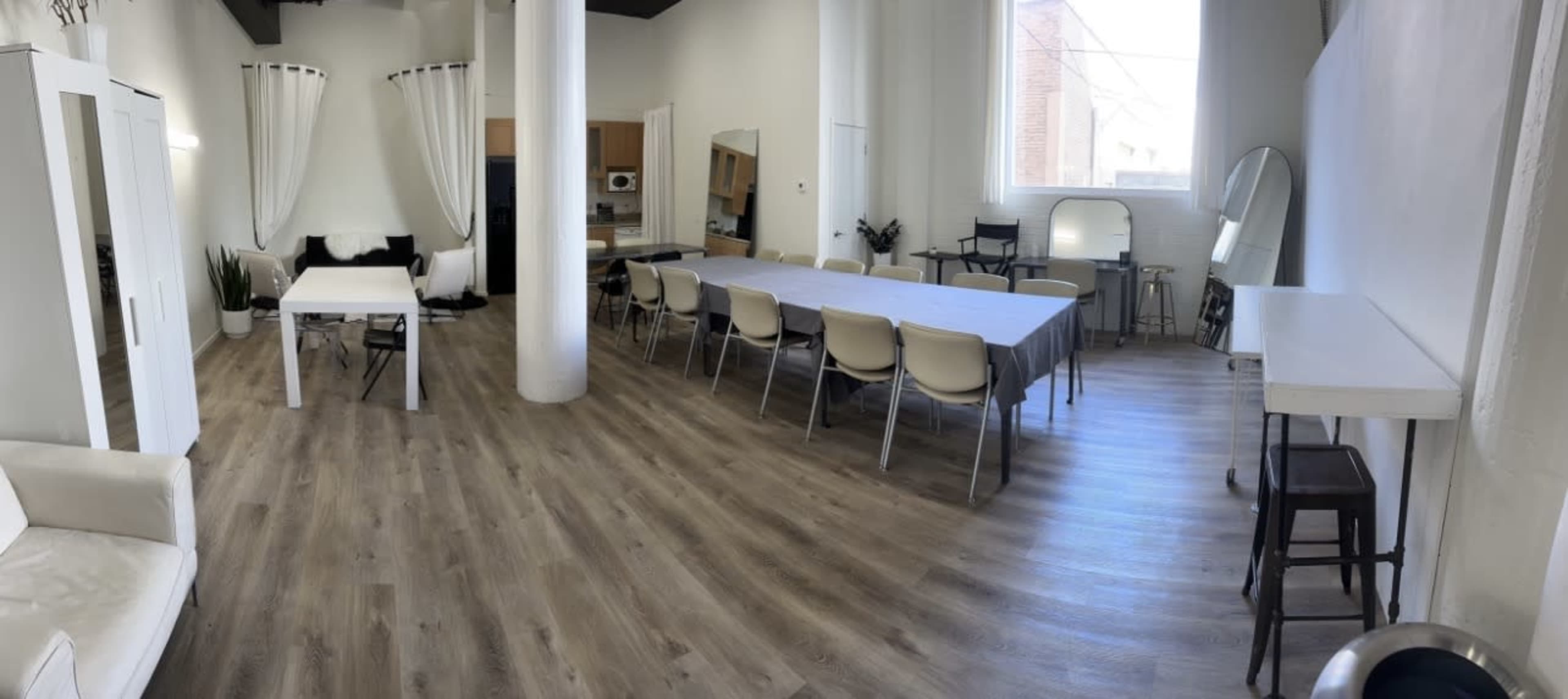 The image shows a spacious, well-lit interior space featuring a long dining table, a smaller table, a kitchen area, and a cozy seating area with white furniture.