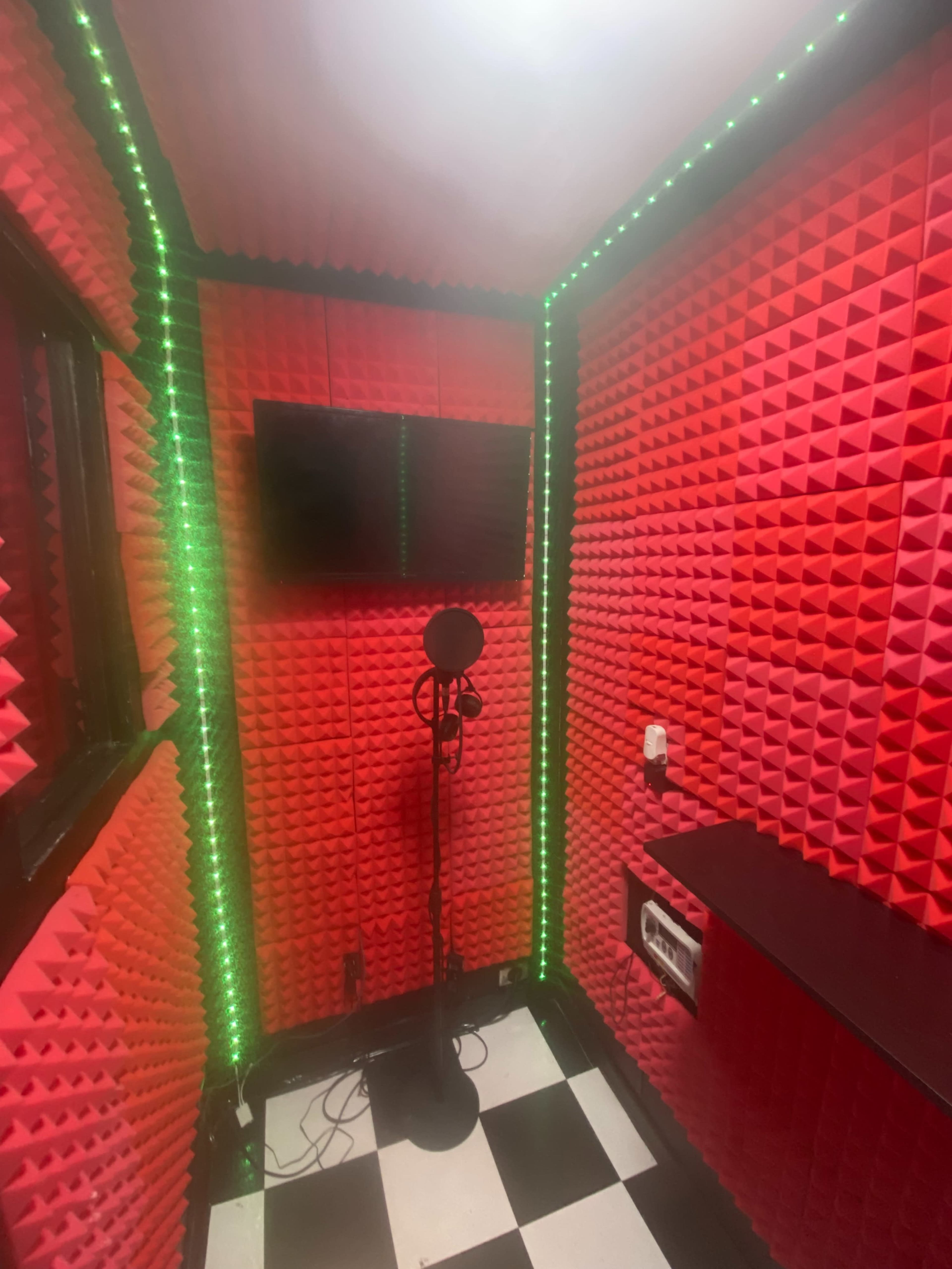 A small recording booth with red acoustic foam walls, a microphone on a stand, and a green LED light along the corners.