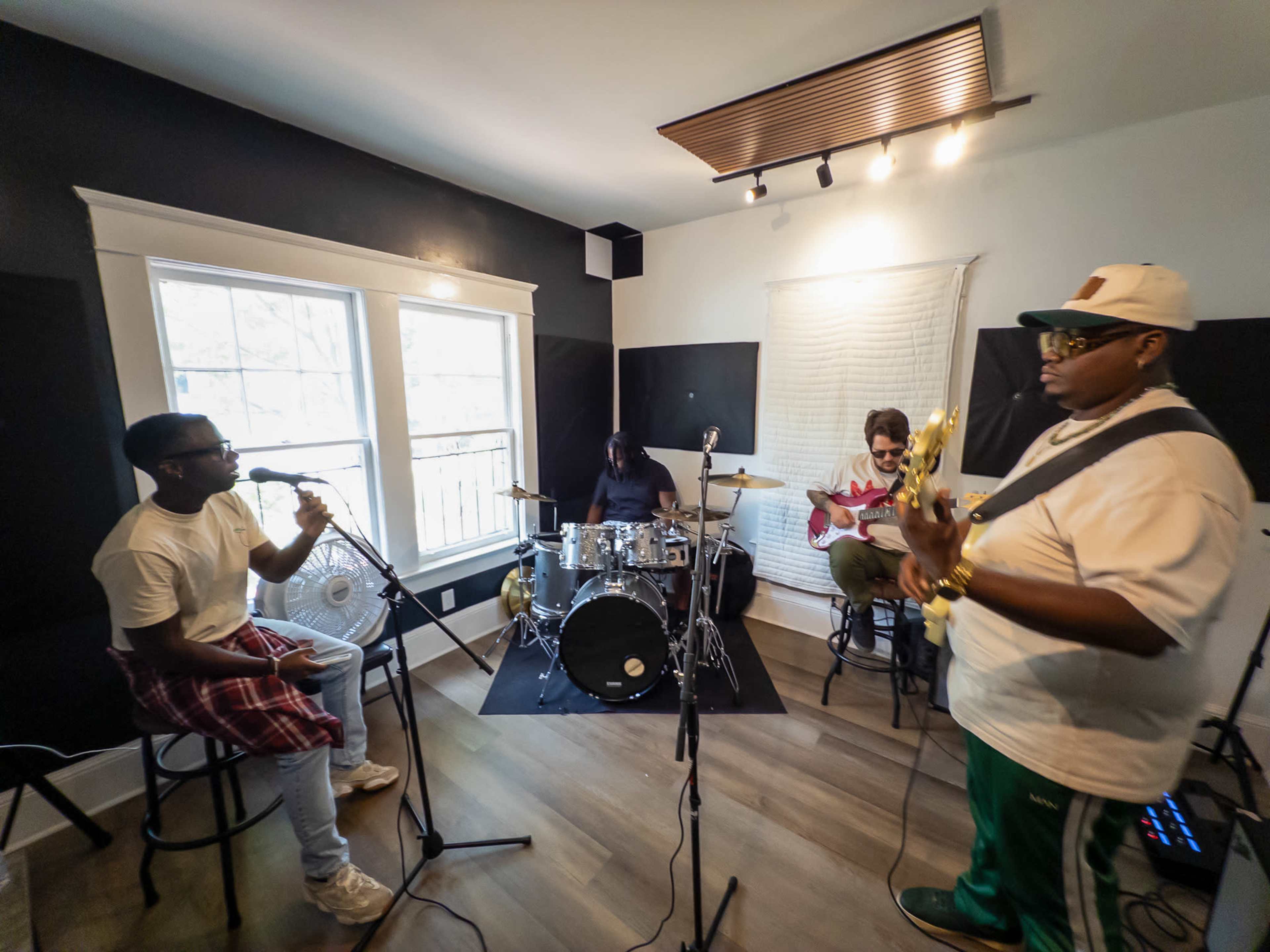 A band of four musicians is rehearsing in a studio, with one singer at a microphone, a drummer, and two guitarists.