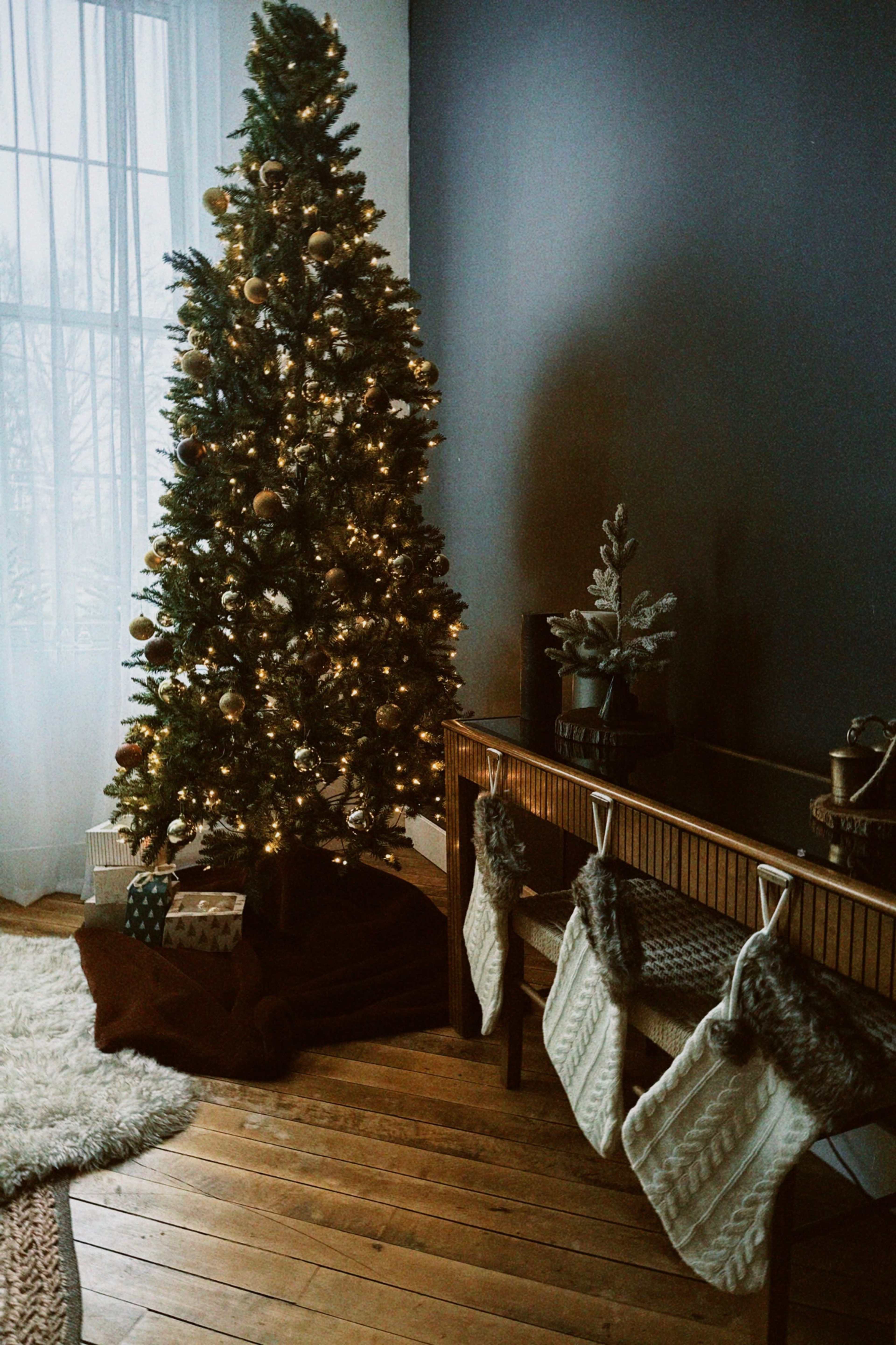 A decorated Christmas tree stands in the corner of a room beside a console table with stockings hanging from it.
