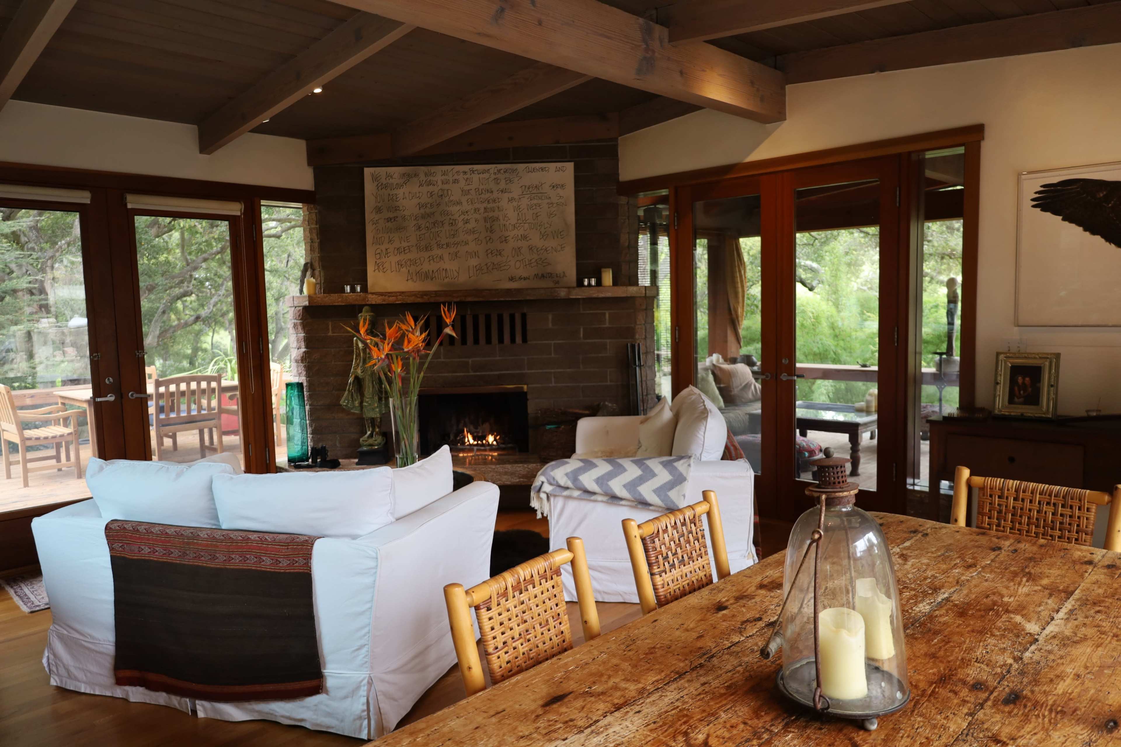 The image shows a cozy living area with a stone fireplace, comfortable seating, and large windows overlooking a garden.