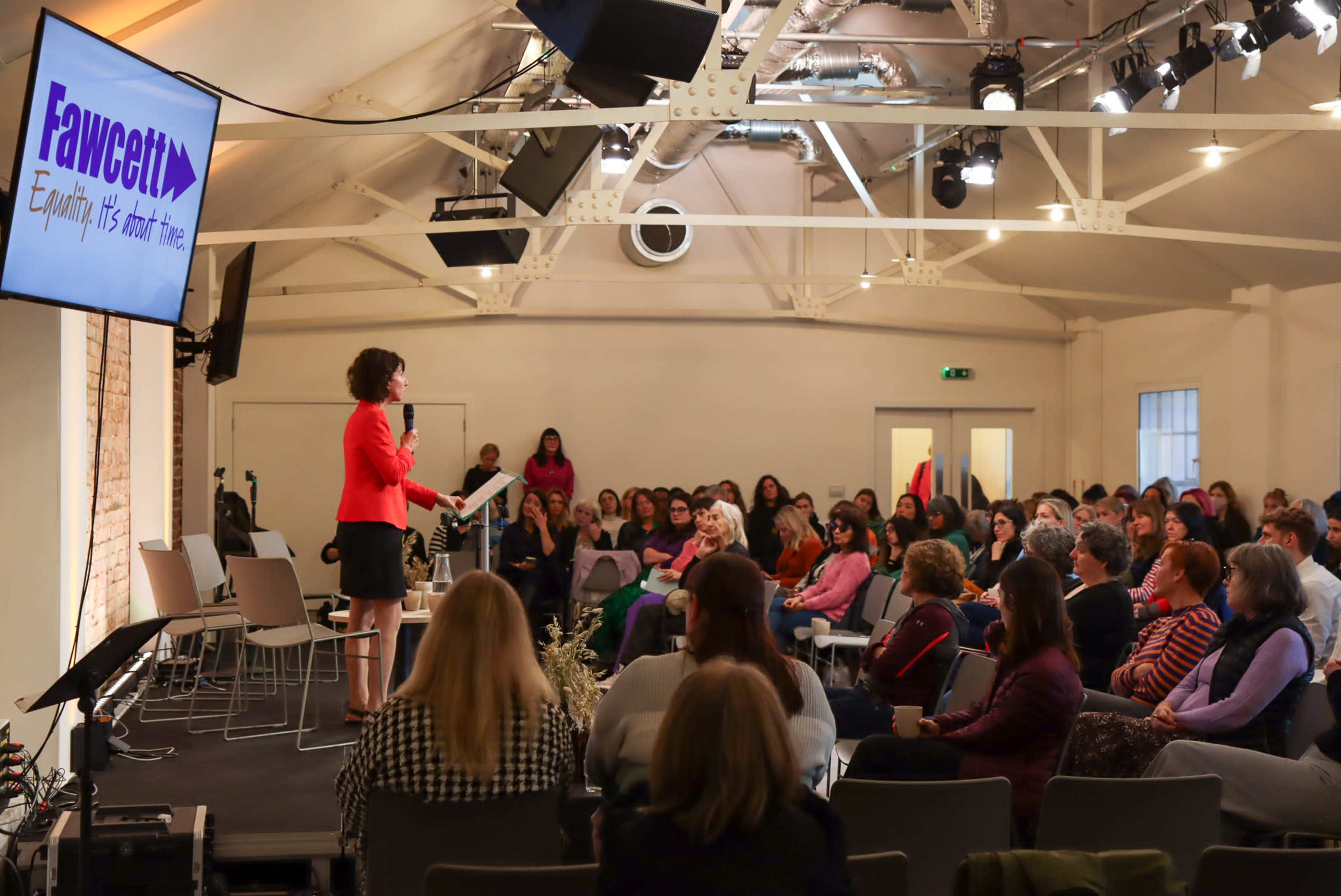 A speaker stands on a stage addressing an attentive audience in a bright, open meeting space.