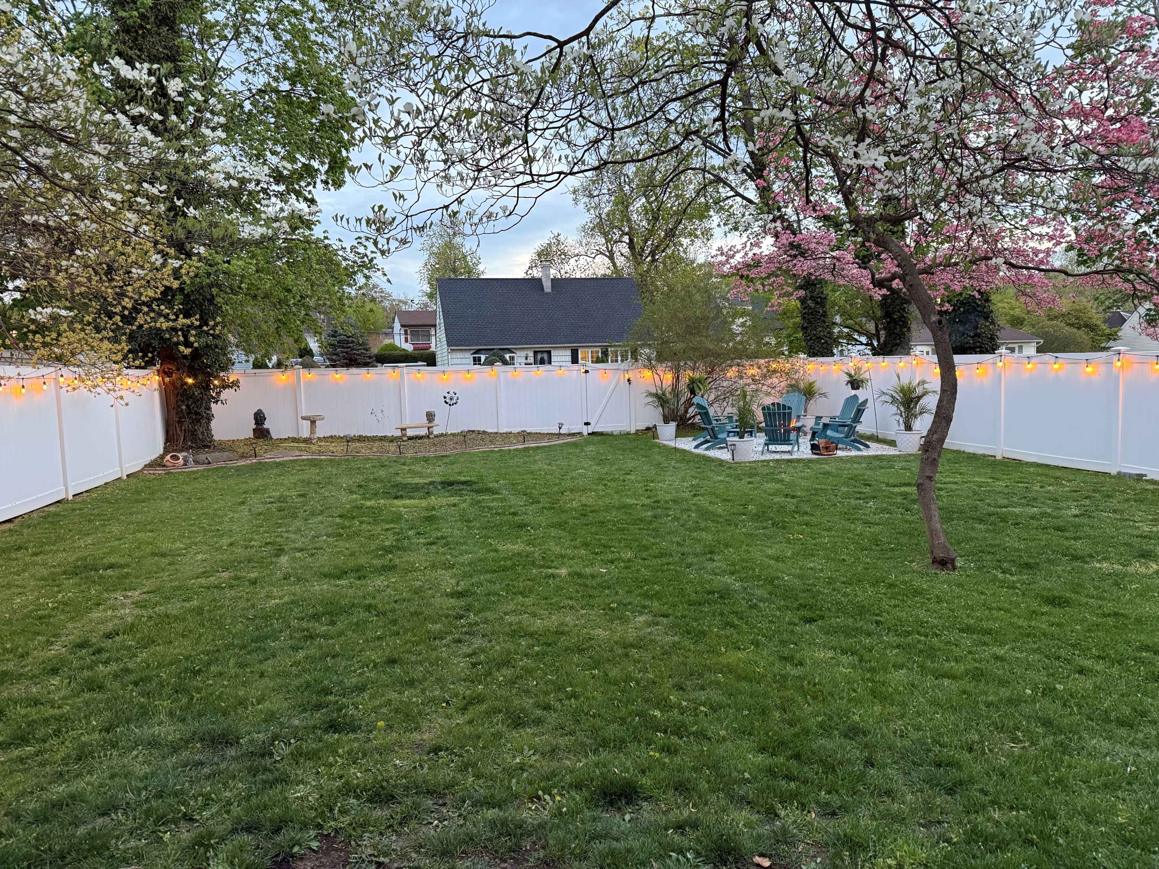 The image shows a spacious backyard with a green lawn, bordered by a white fence and decorated with string lights, featuring a seating area with chairs and a small patio next to a tree with blooming flowers.