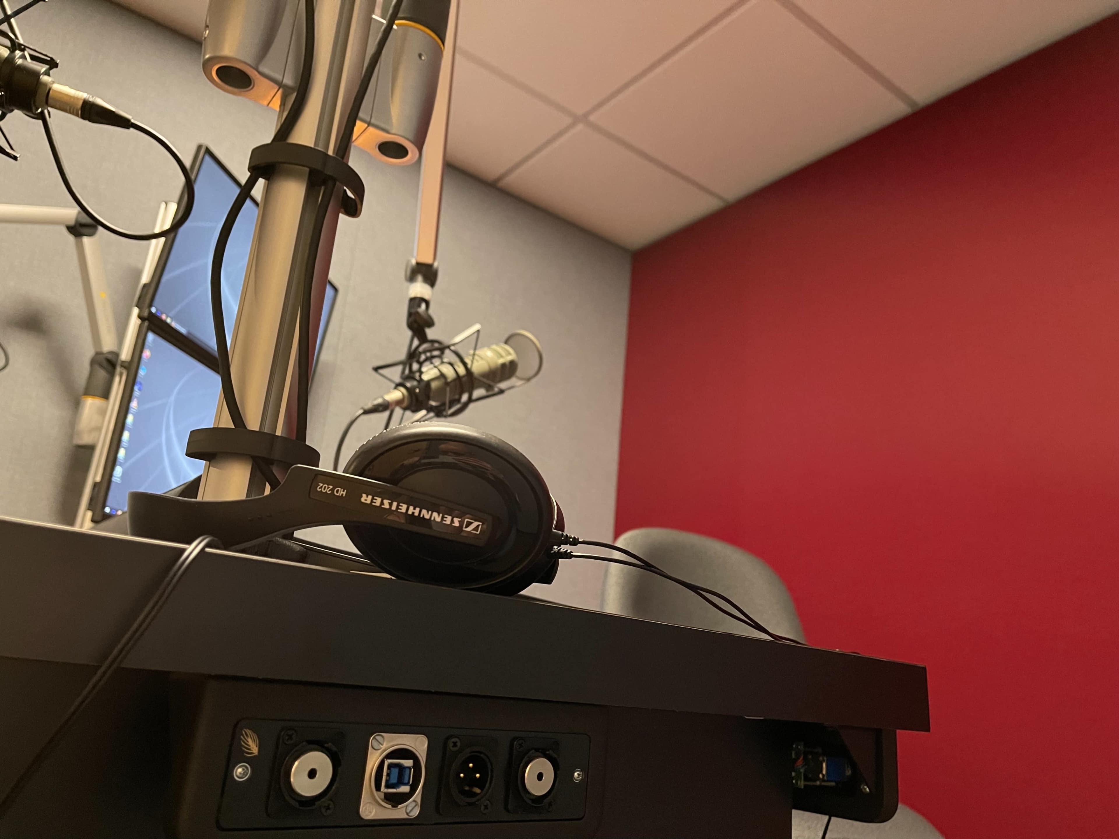 A microphone and headphones sit on a table in a recording studio with a red accent wall.