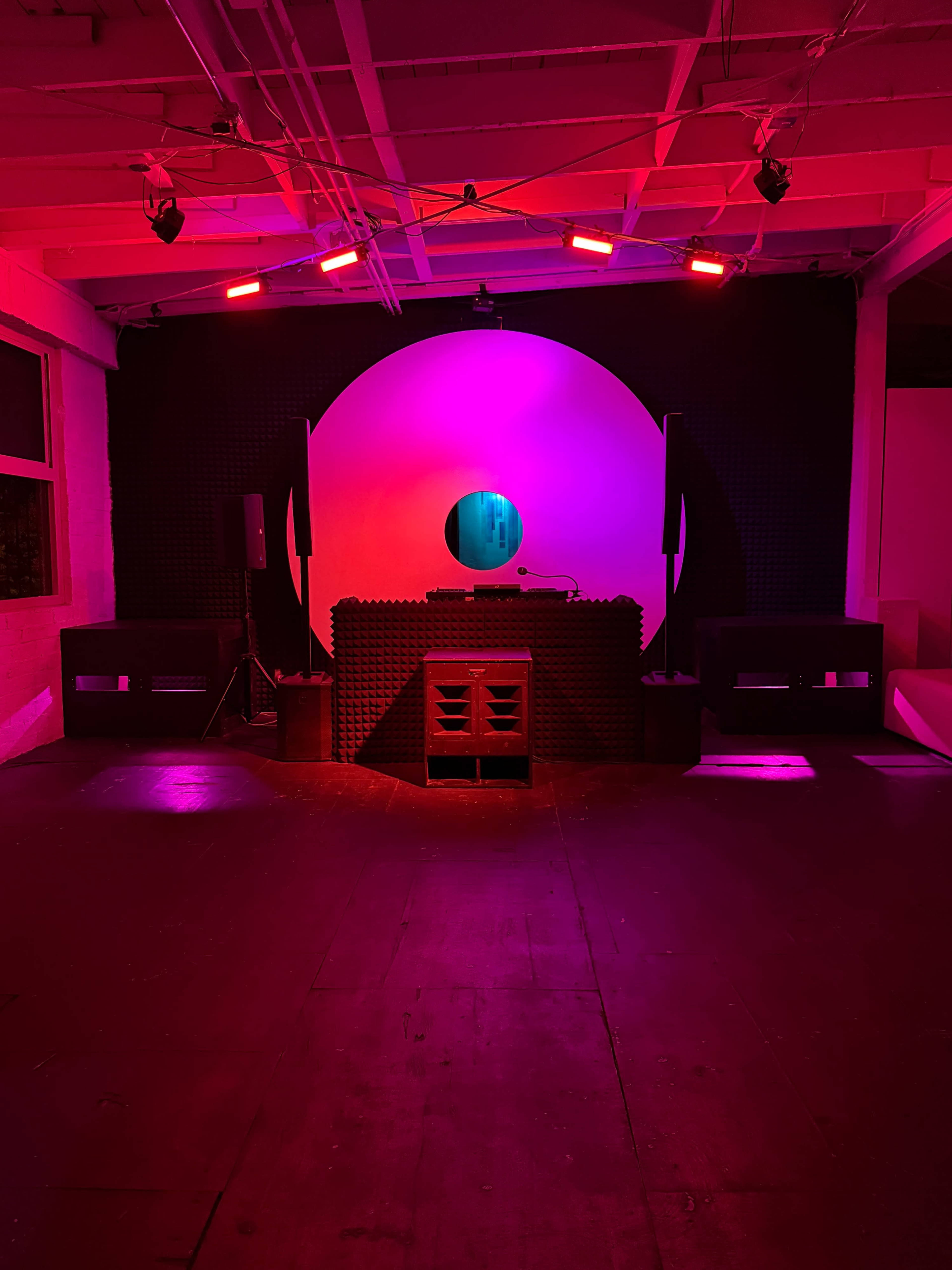 The image shows a dimly lit space with a DJ booth featuring a circular backdrop, illuminated by red and purple lights.