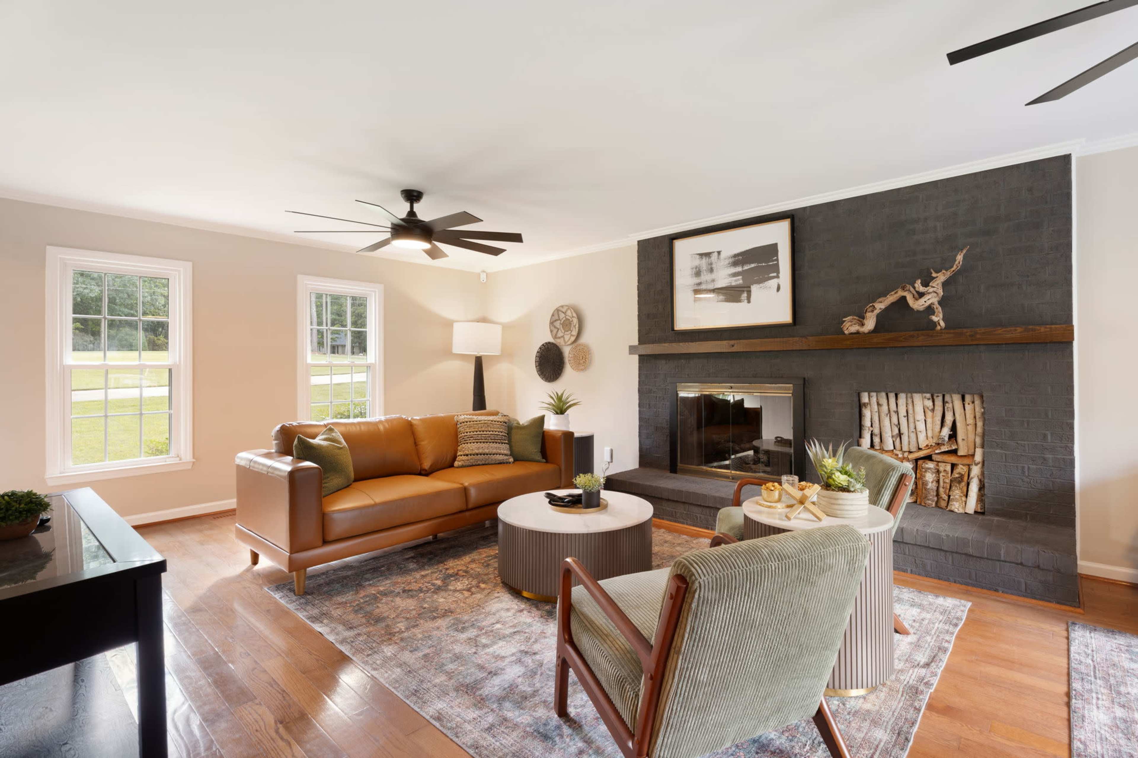 The image shows a cozy living room featuring a brown leather sofa, a round coffee table with snacks, a fireplace with decorative logs, and large windows allowing natural light to enter.