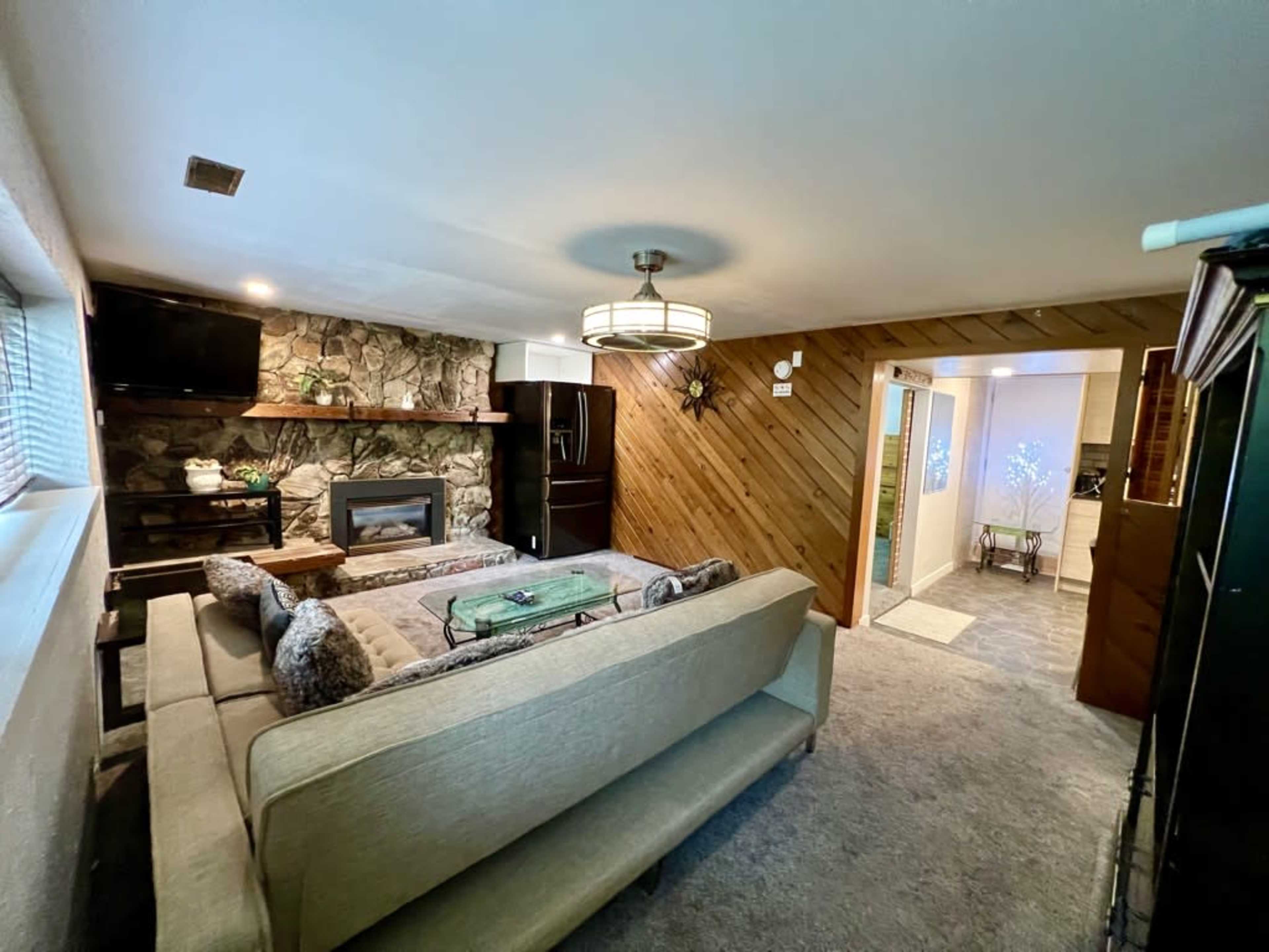 A cozy living room features a sectional sofa, a stone fireplace, and a television mounted on the wall, with a doorway leading to a separate area.