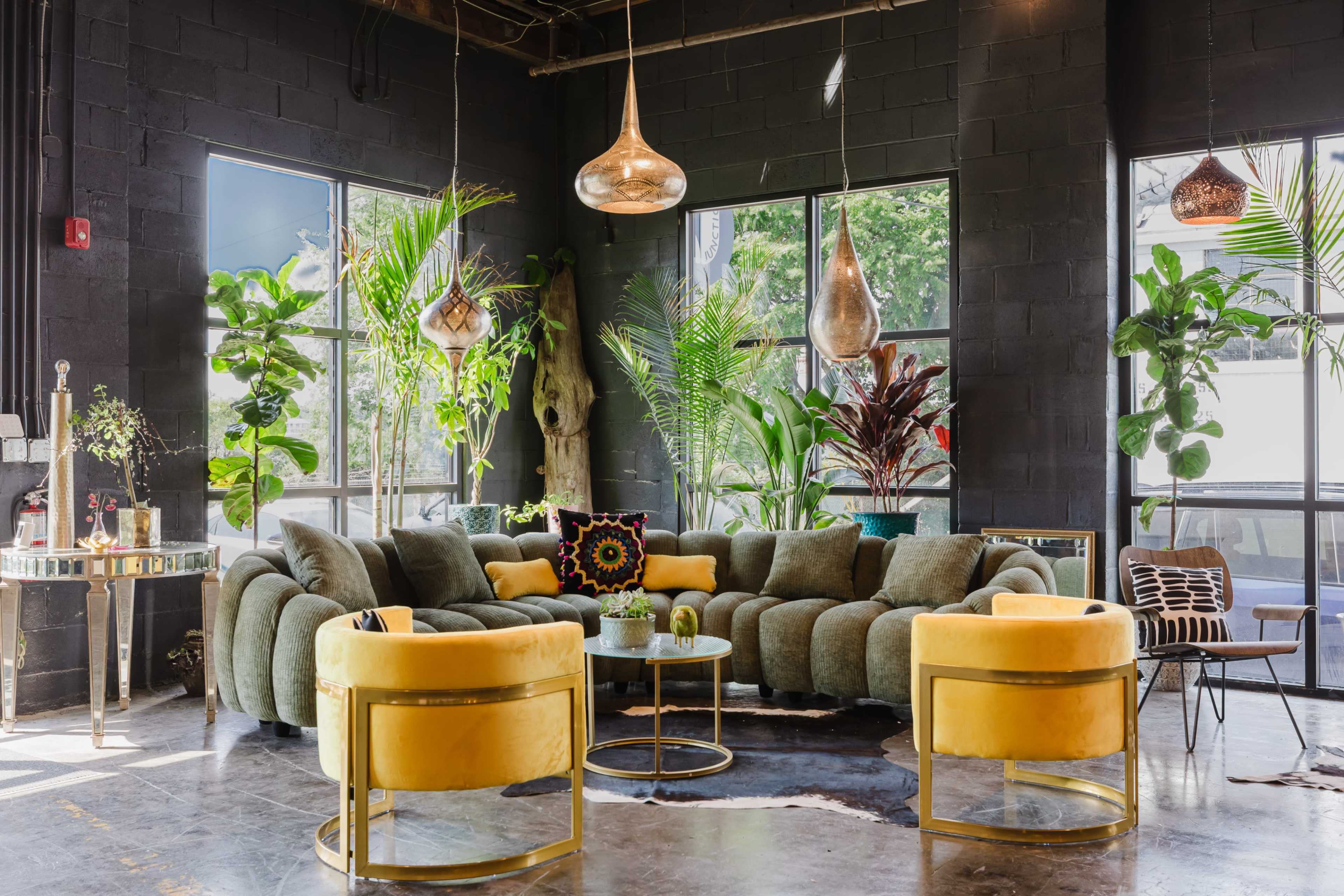The image shows a modern lounge area featuring a large curved gray sofa and yellow accent chairs, surrounded by various indoor plants and large windows allowing natural light.