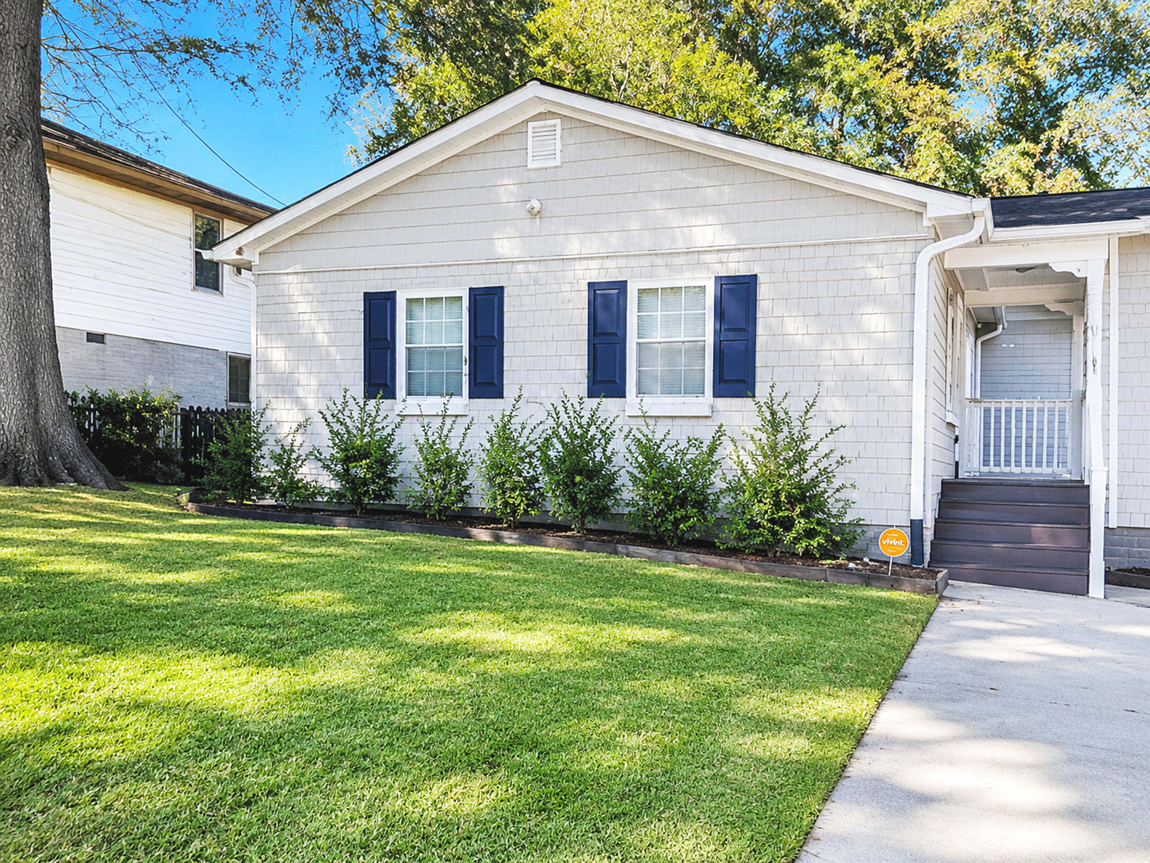 A light-colored house with blue shutters sits on a well-manicured lawn, bordered by a row of shrubs.