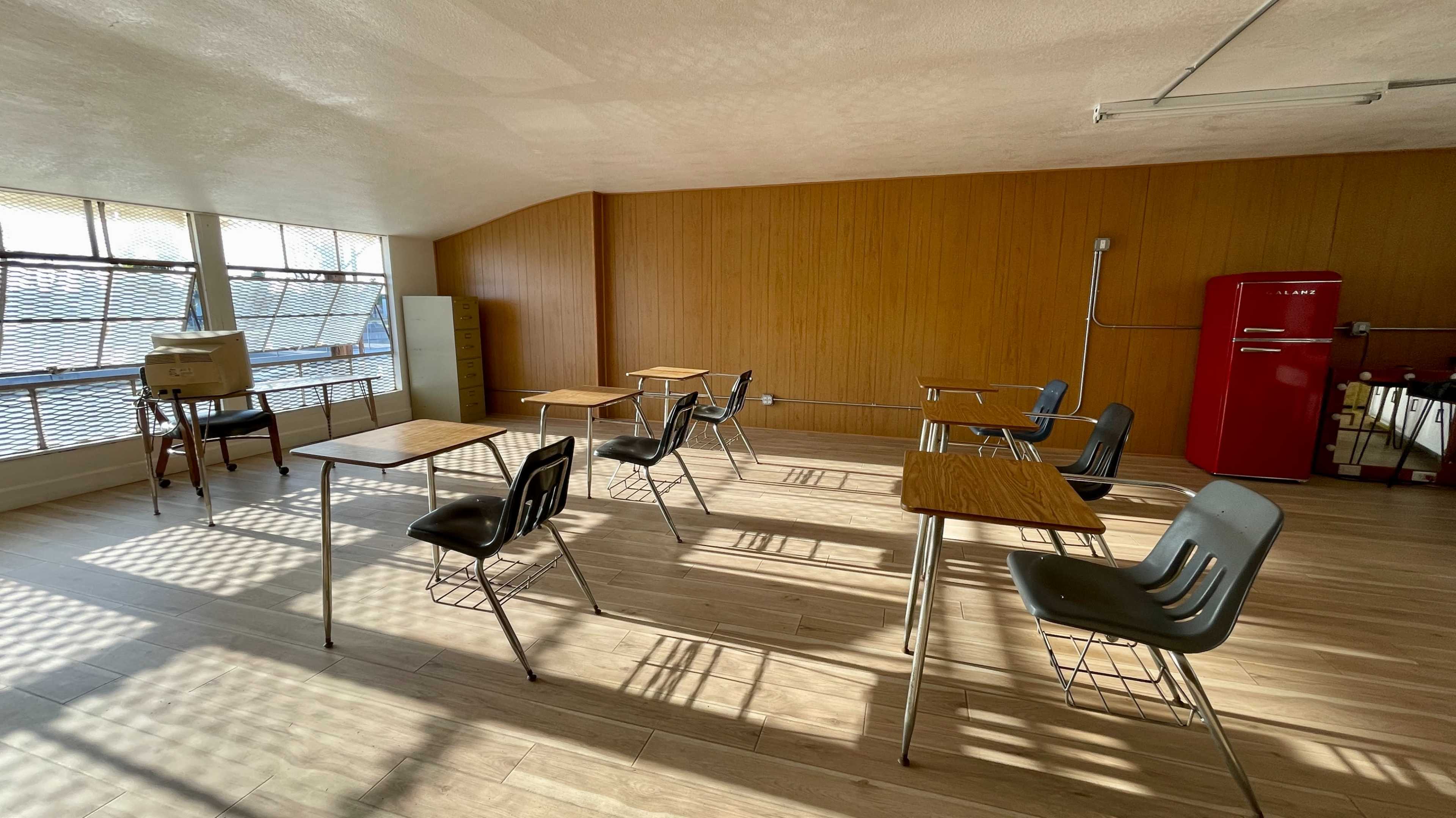 A brightly lit, empty classroom with wooden walls, several desks arranged with chairs, and a red refrigerator in the corner.