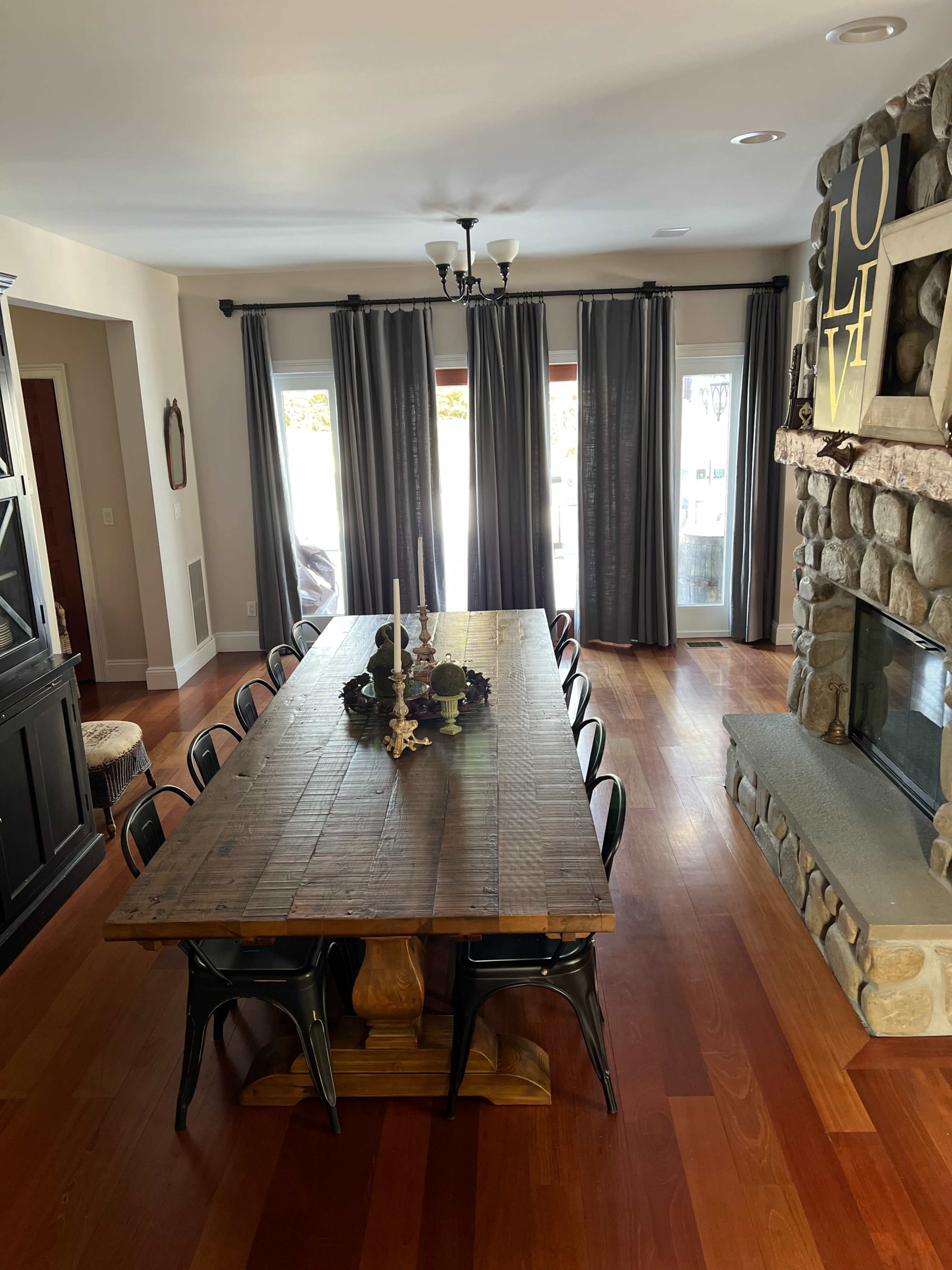 A large wooden dining table with black chairs is centered in a brightly lit room featuring curtains, a stone fireplace, and a view of the outdoors.