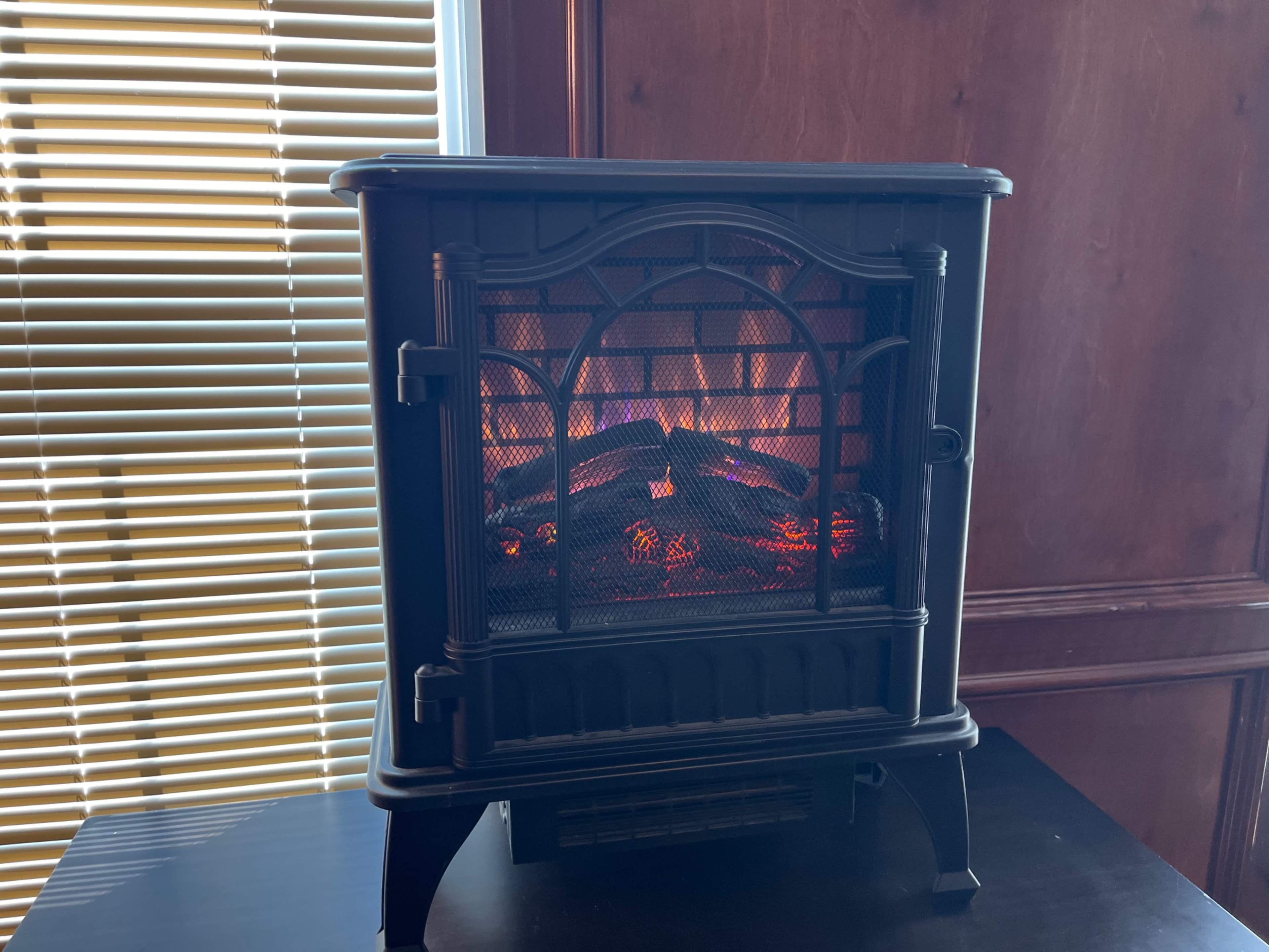 A black electric fireplace sits on a table, emitting a warm glow against a backdrop of wooden panels and window blinds.