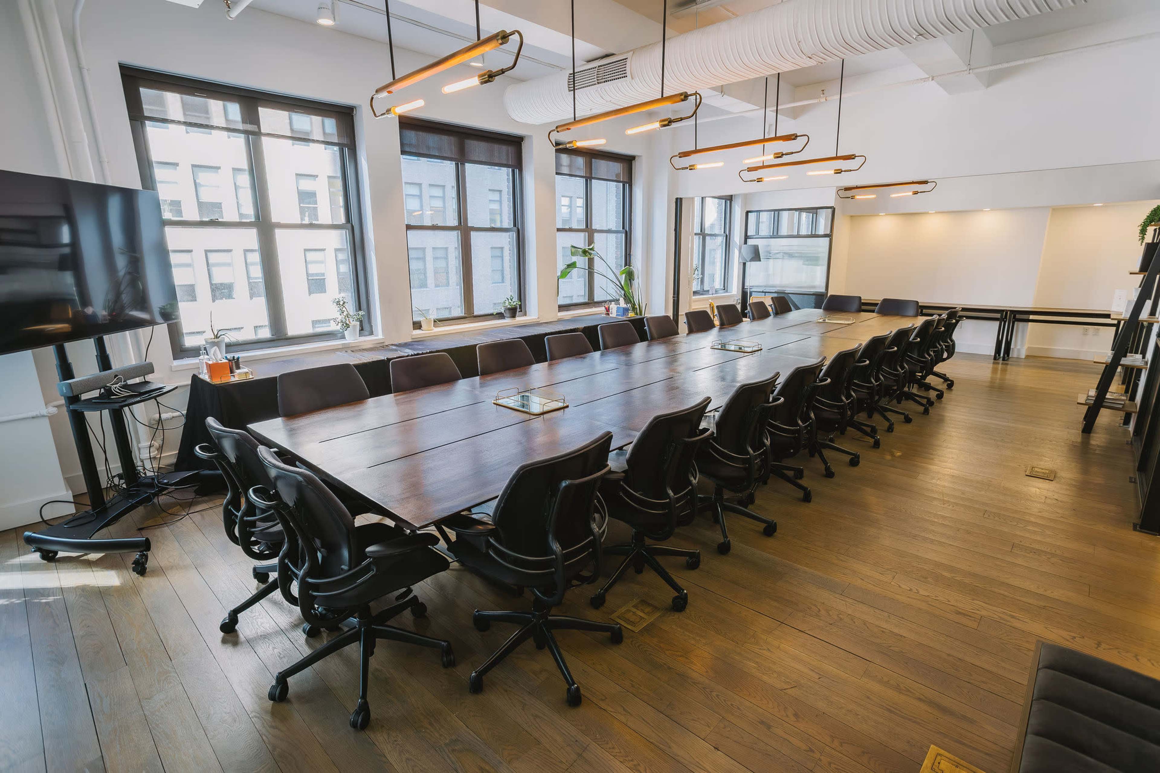 The image shows a large conference room with a long wooden table surrounded by black ergonomic chairs and large windows allowing natural light to enter.