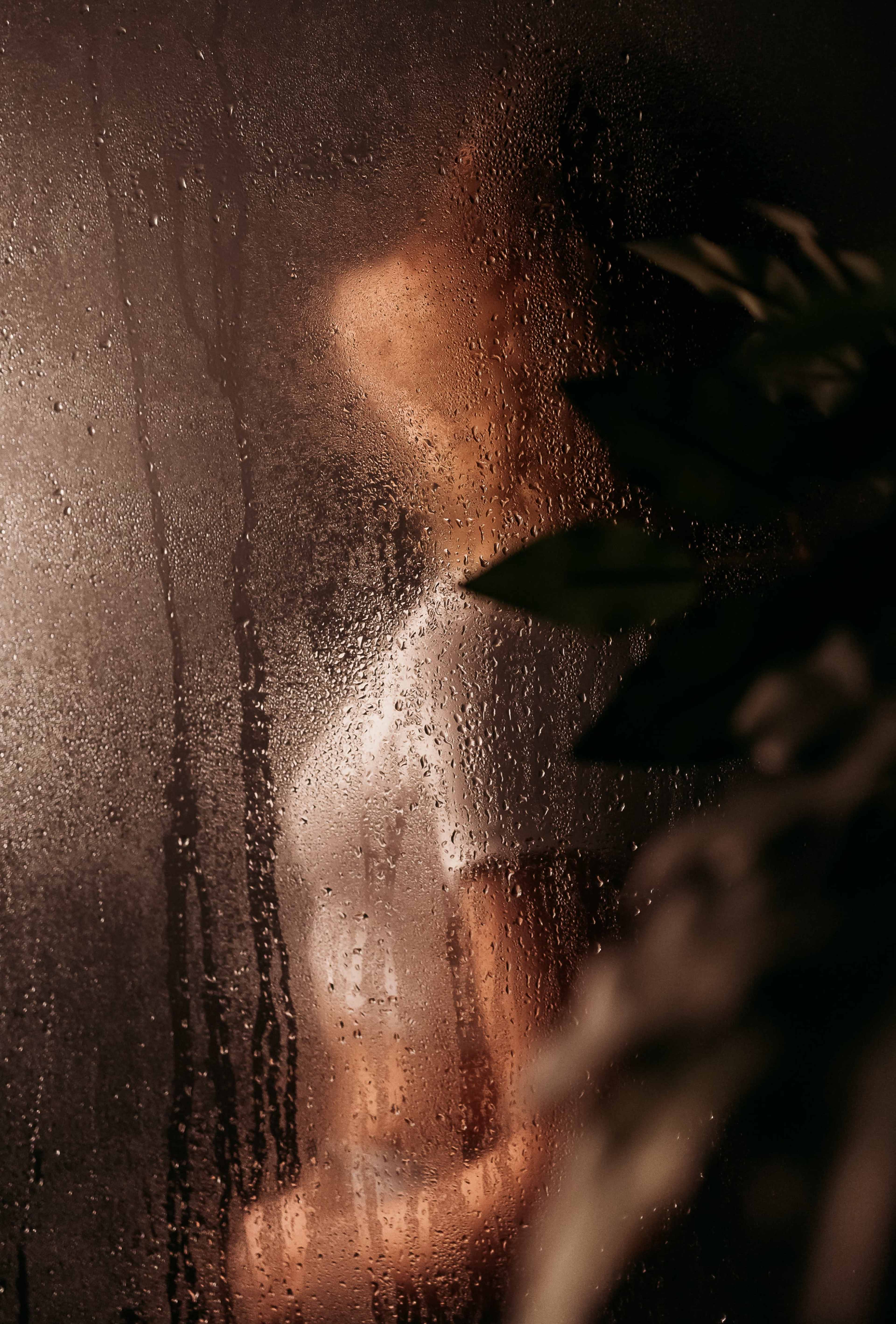 A person stands behind a fogged glass shower door, with water droplets creating a misty effect.