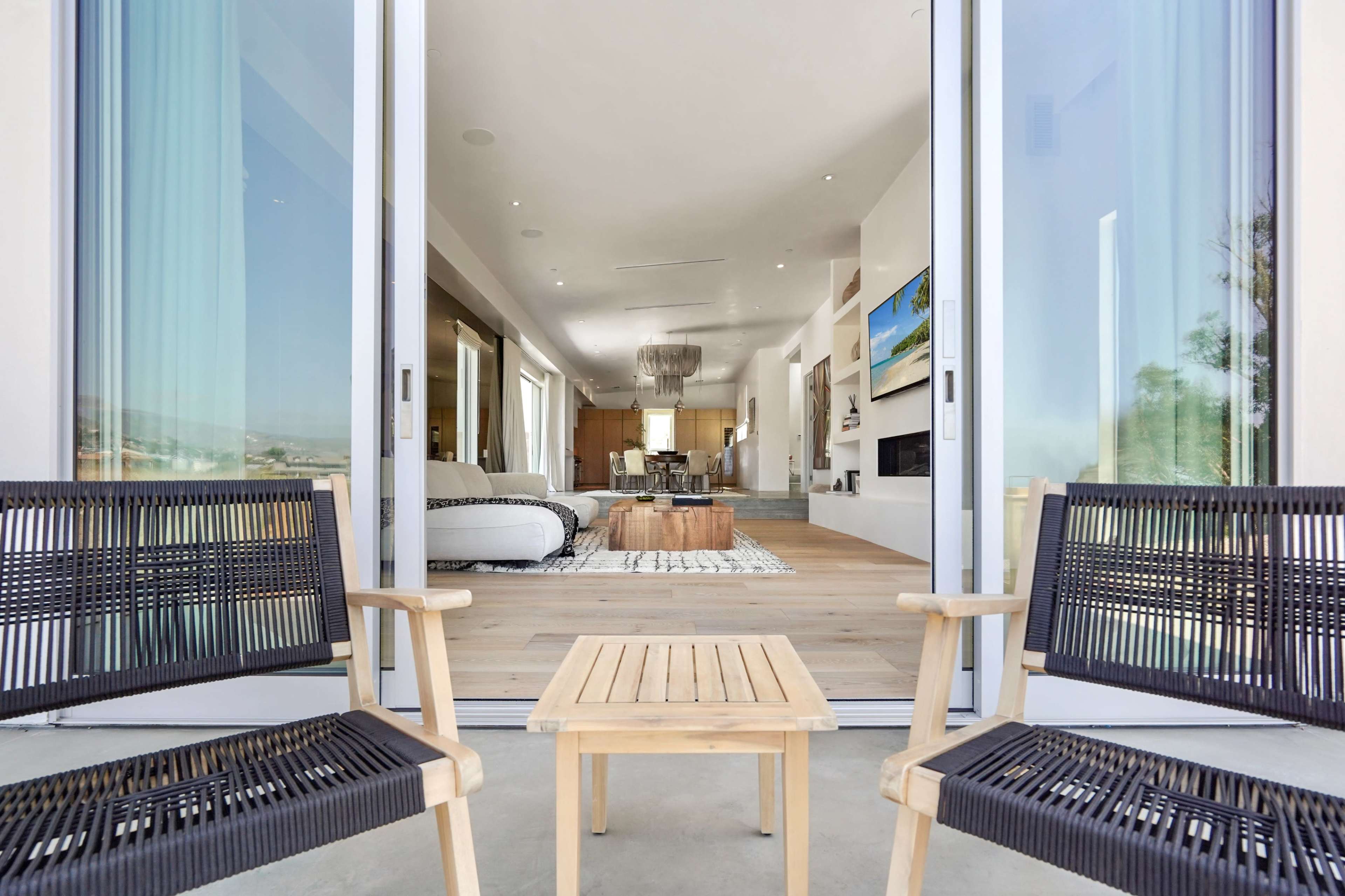 The image shows two chairs and a small table on a patio, overlooking a modern living space with a view of the interior.