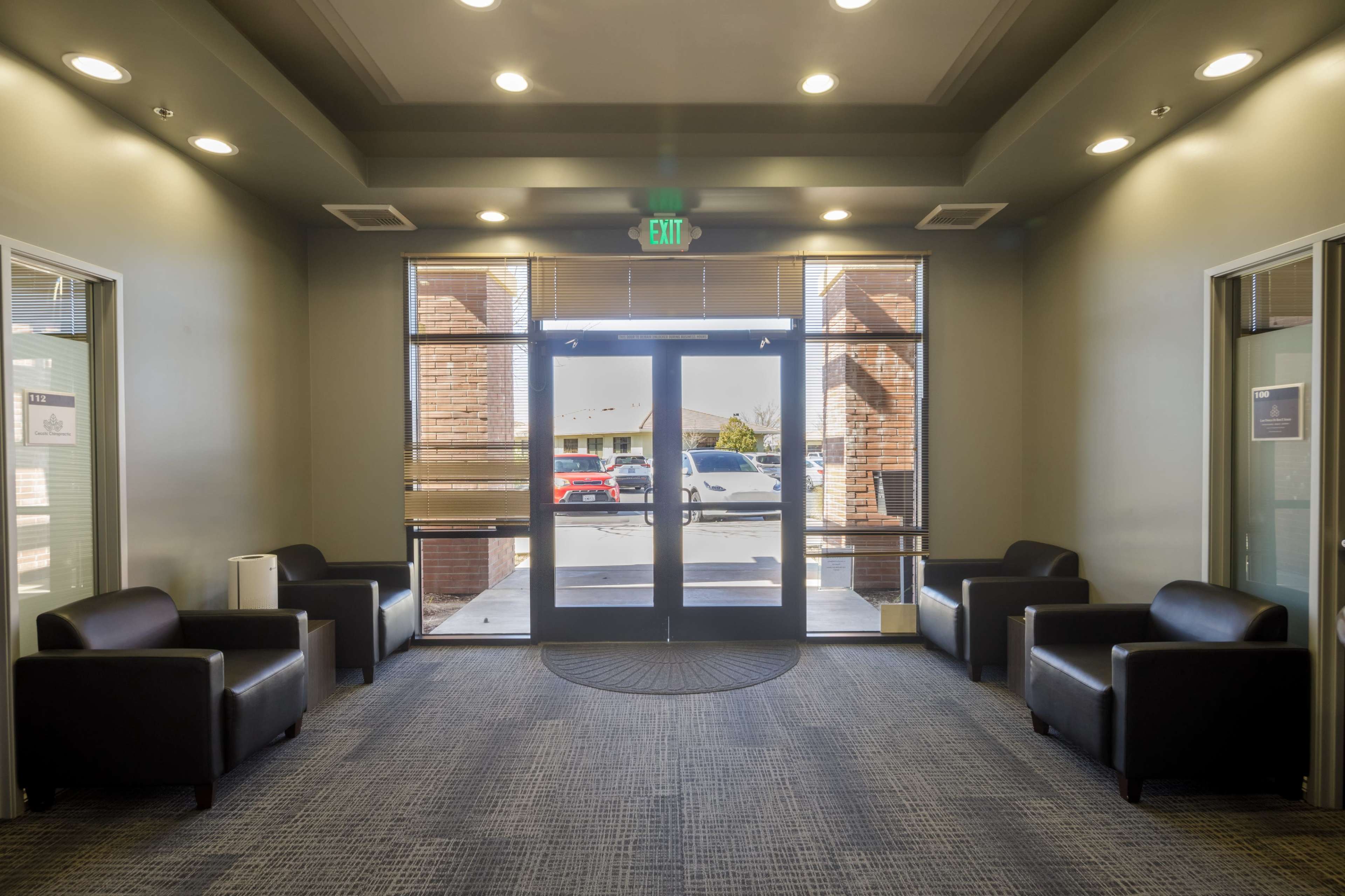 The image shows a modern waiting area with four black chairs arranged around a circular carpet, facing a set of double glass doors that lead outside.